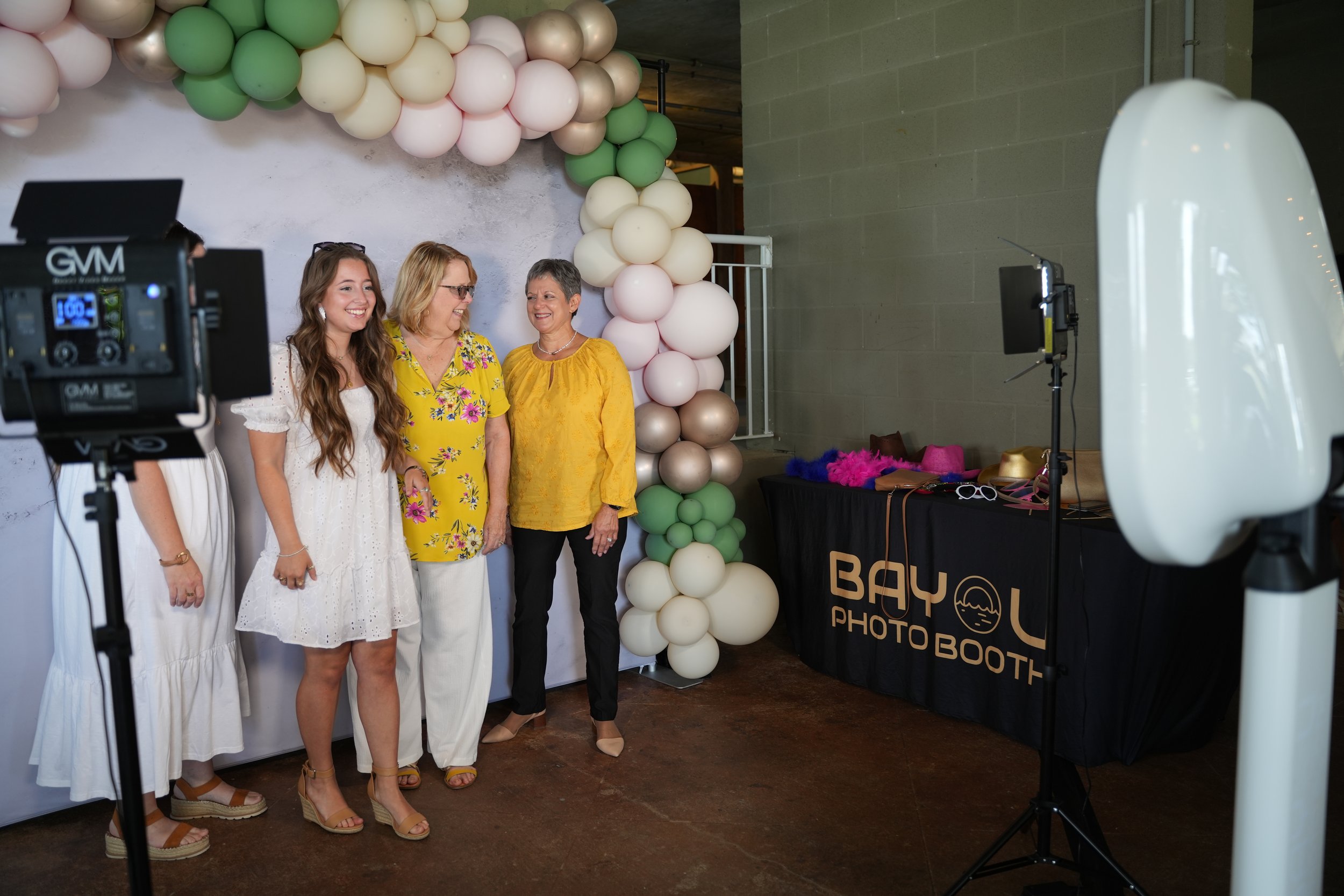 Three women and a girl pose for a photo at a photo booth with a balloon arch in the background. The photo booth has a black table with props, and a camera setup is visible in the foreground.