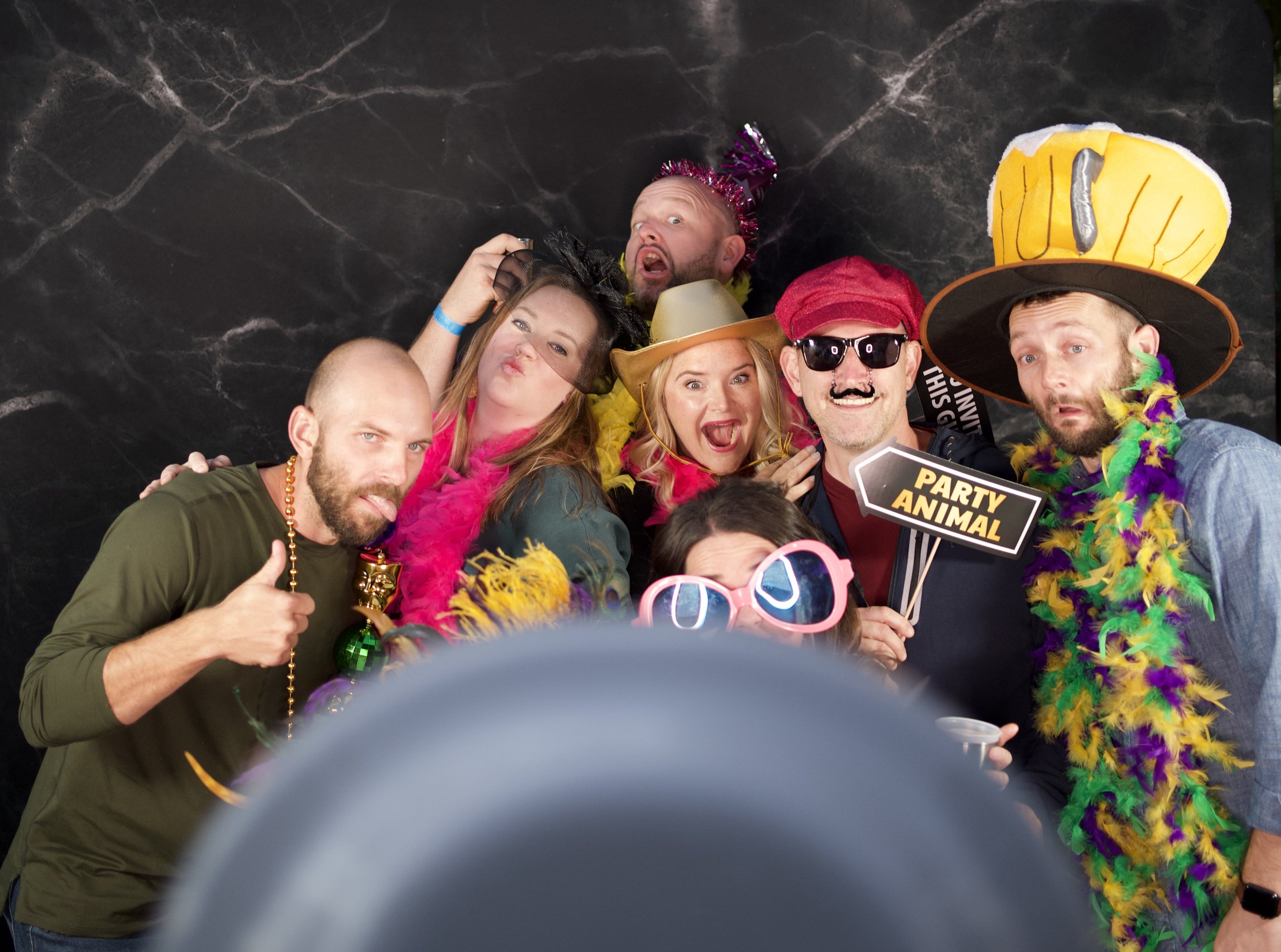 Group of people at a party dressed in colorful costumes and accessories, with some holding props like a "Party Animal" sign and wearing novelty hats and glasses against a black backdrop.