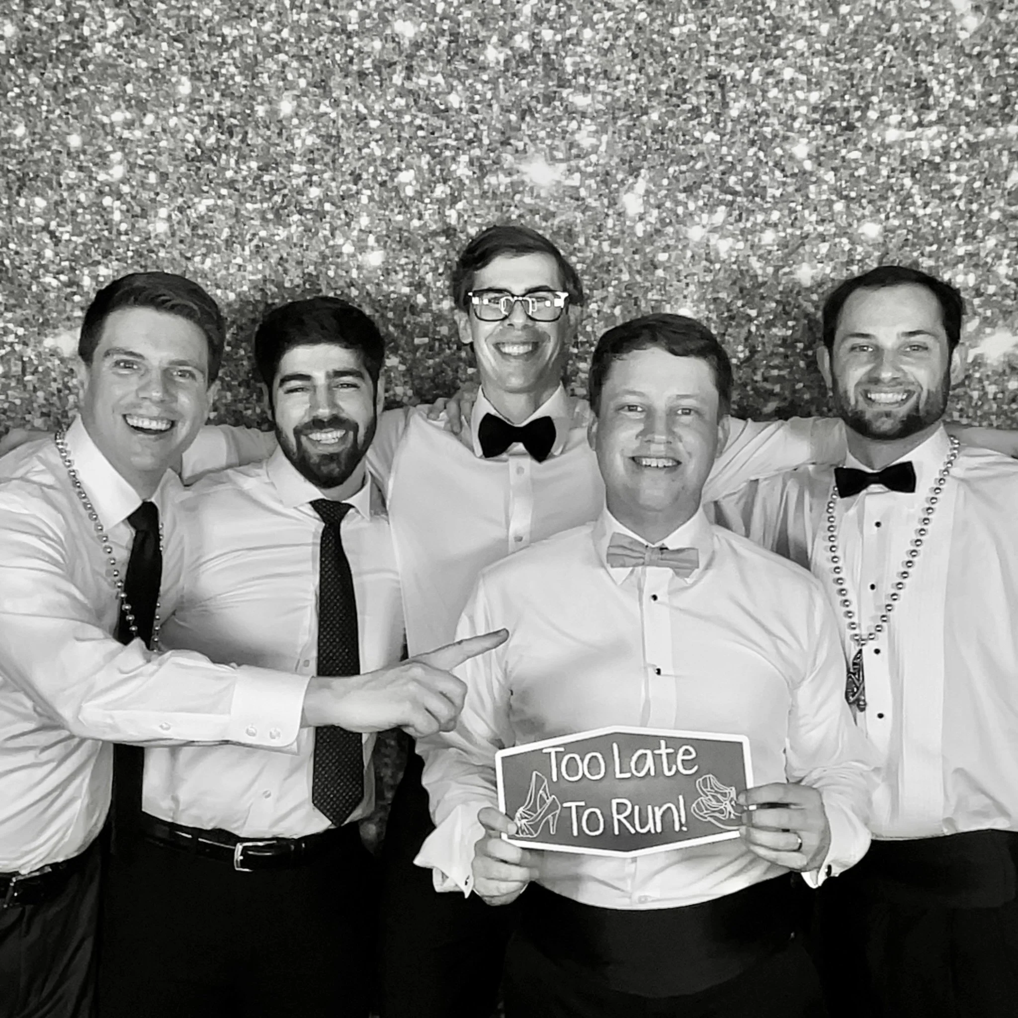 Group of six men dressed in white shirts and ties or bow ties, posing together with smiles against a glittery backdrop. One man in front is holding a sign that reads 'Too Late To Run' and points at it.