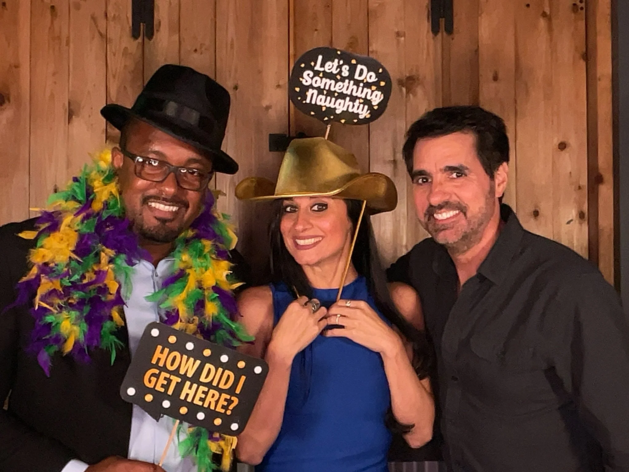 Three people smiling at a photo booth, dressed in fun costumes with props, holding signs that read 'How did I get here?' and 'Let's do something naughty,' with a wooden wall background.