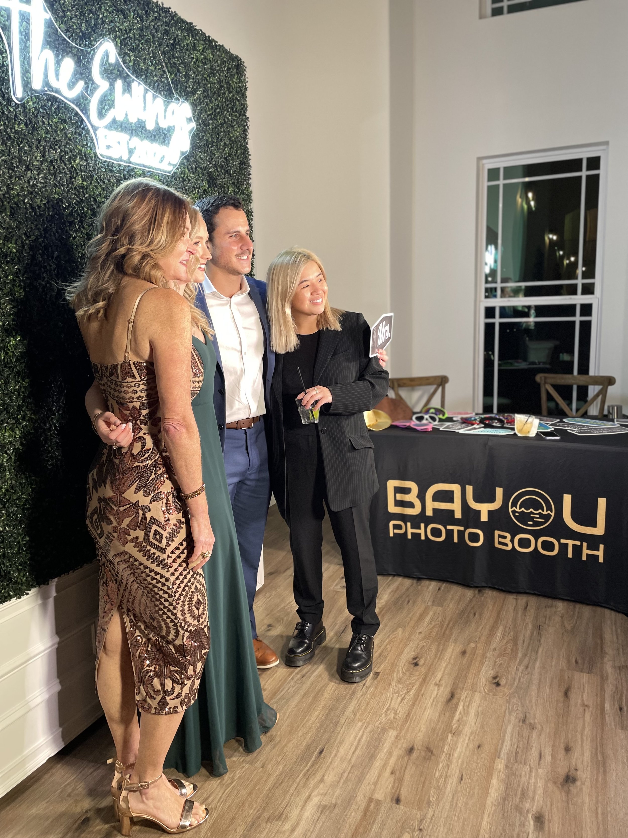 Four people posing for a photo at a photo booth with a sign that reads 'The Elmen' in cursive neon letters. They are standing in front of a green hedge wall, and there is a black table with photography props and a sign that says 'BAYOU PHOTO BOOTH.'