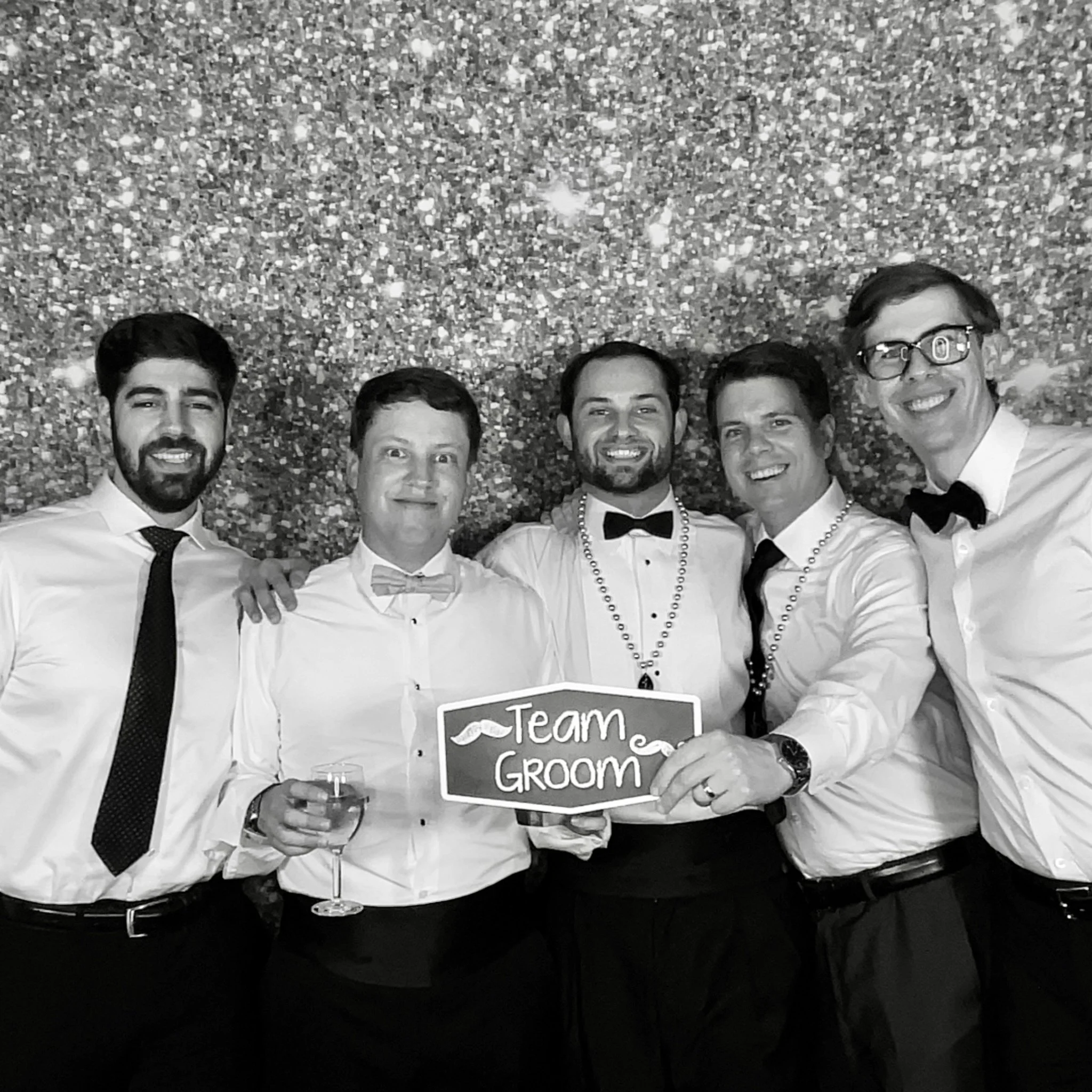 Five men dressed in formal attire, smiling in front of a glittery backdrop. One man in the center holds a sign that says 'Team Groom' and a glass of wine, suggesting it is a wedding or bachelor party celebration.