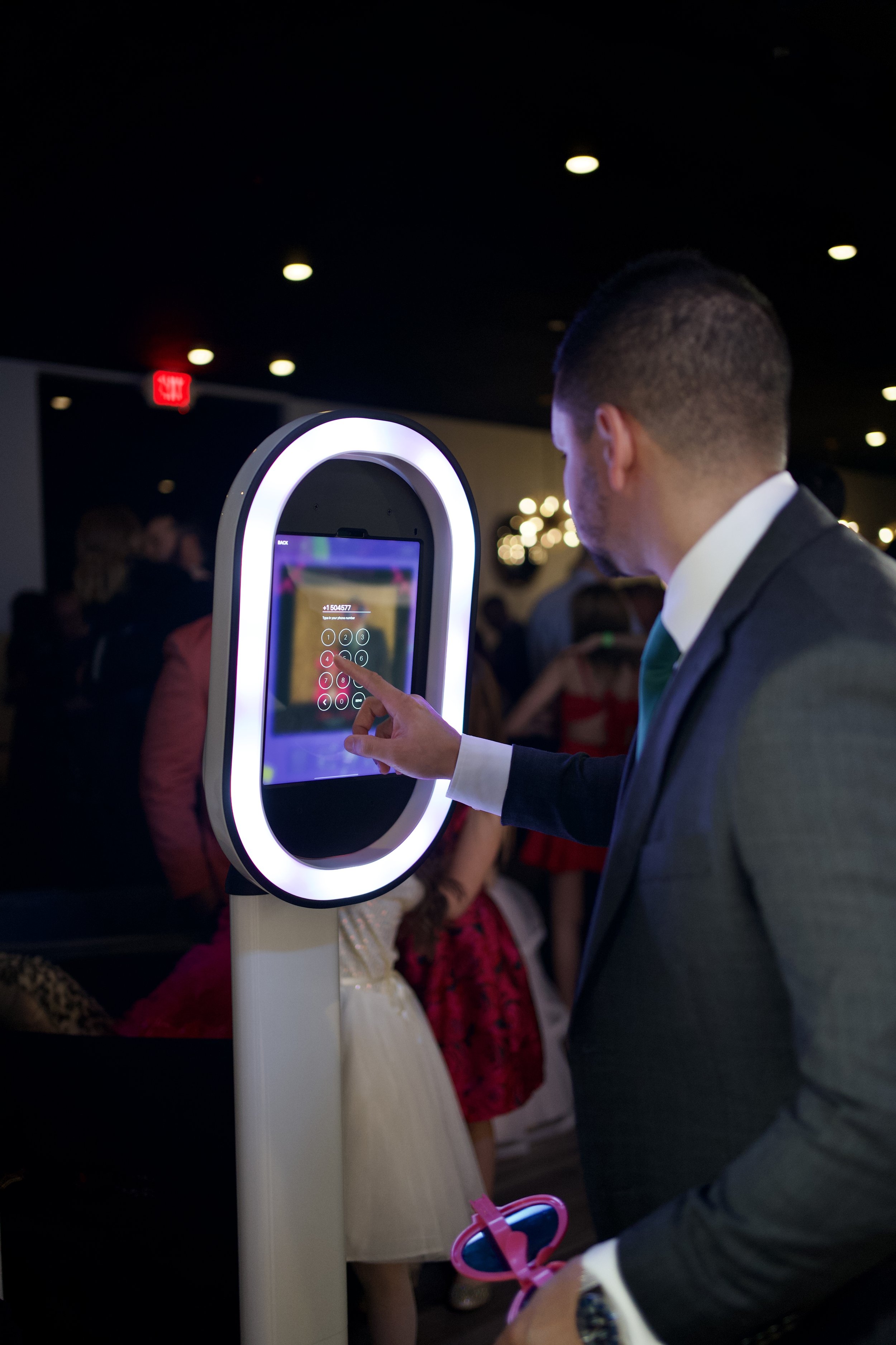 Man dressed in a suit using a touchscreen kiosk at a social event with people in fancy attire in the background.