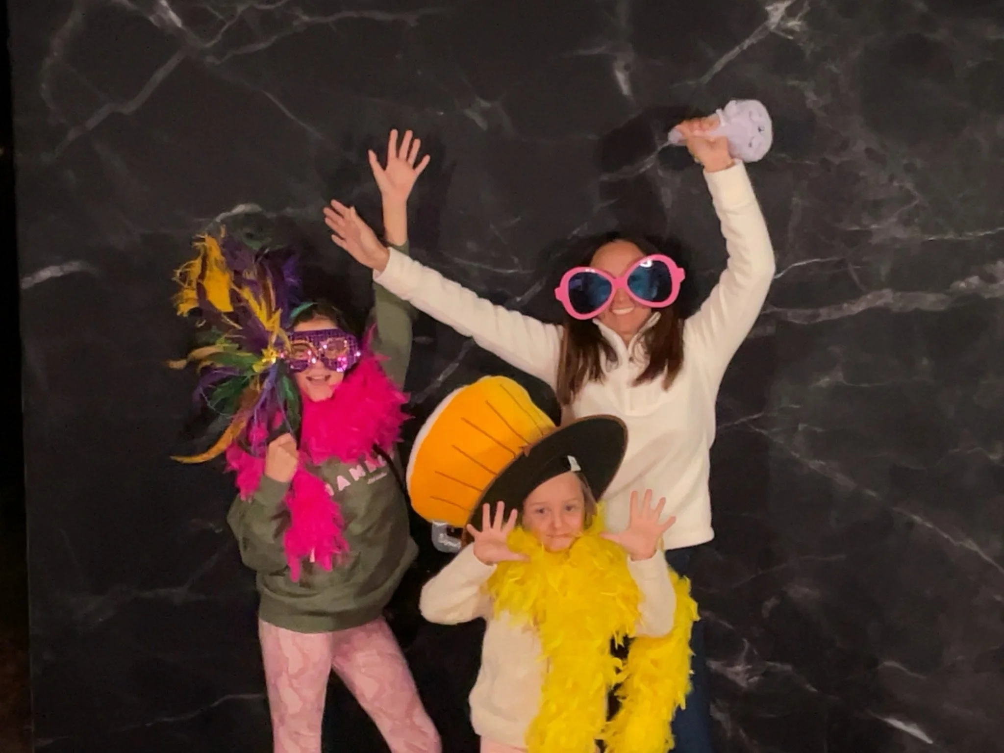 Three people, two children and one adult, wearing colorful fun costumes and accessories, posing playfully against a dark textured background.