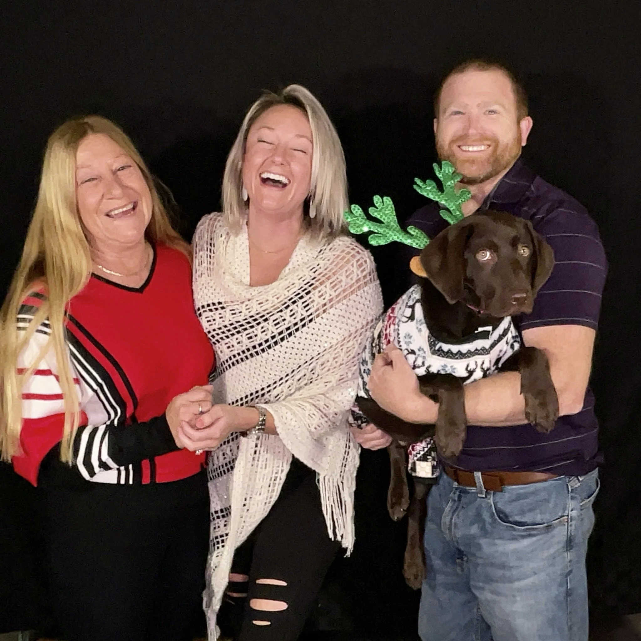Three women and one man smiling, the man is holding a puppy wearing reindeer antlers and a holiday sweater, all against a black background.