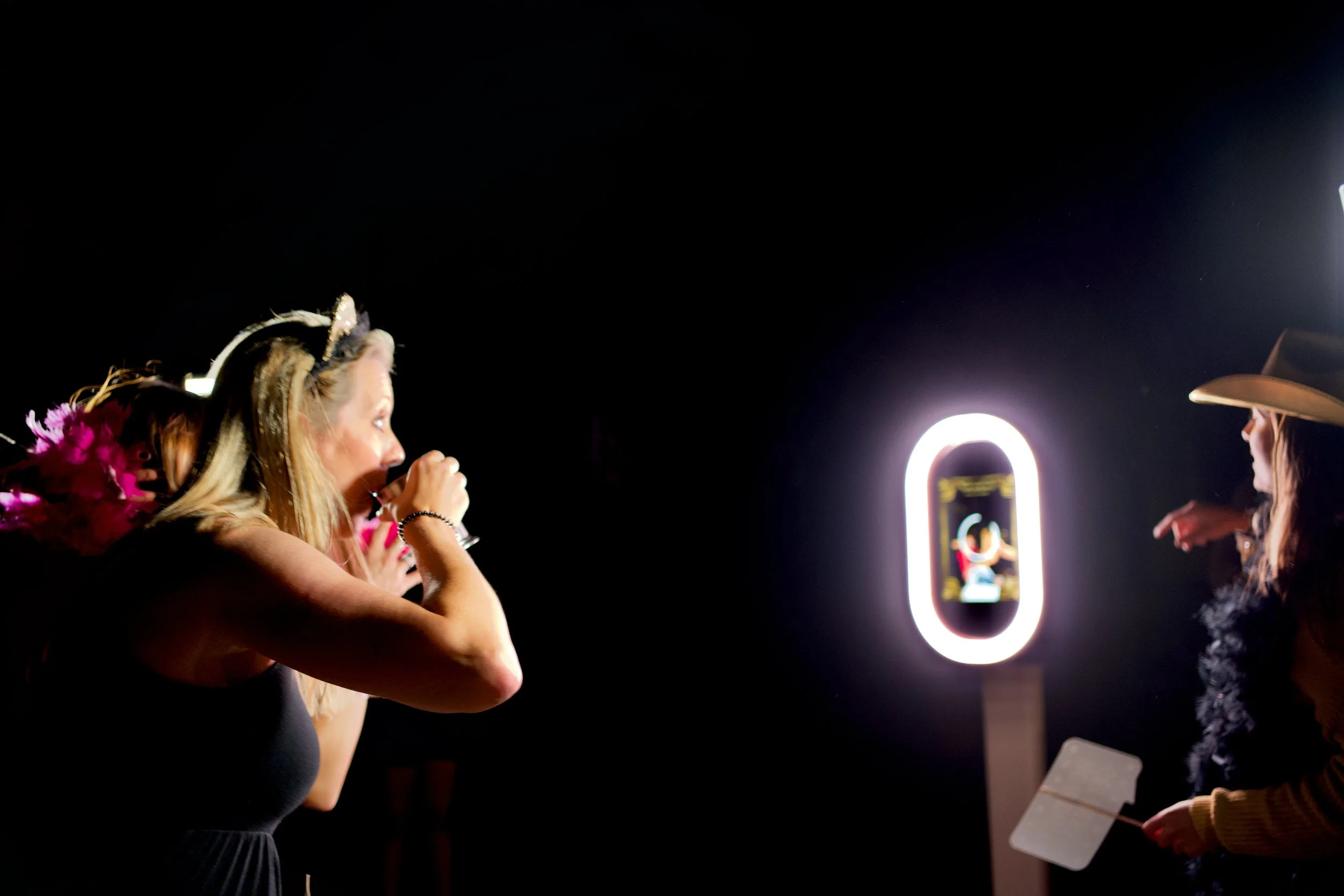 A woman with a headband and pink feathered accessory blows bubbles at night, illuminated by neon signs in the background.