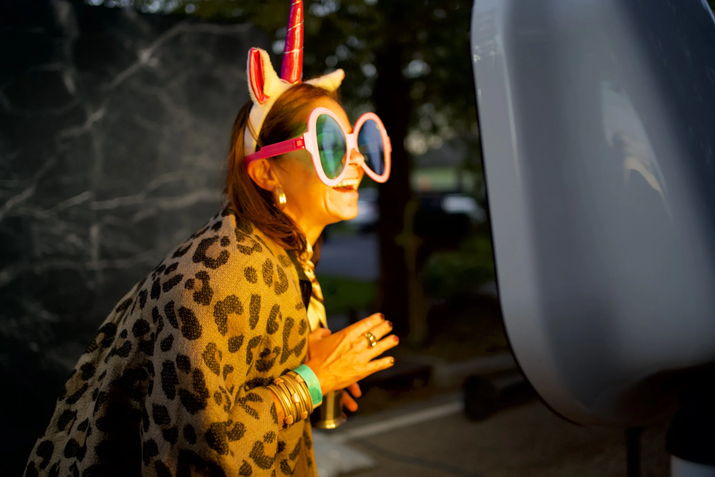 A woman wearing a playful unicorn horn and large sunglasses, with a zebra print jacket and multiple bracelets, appears to be looking at a mirror or a shiny surface while smiling outdoors during the daytime.