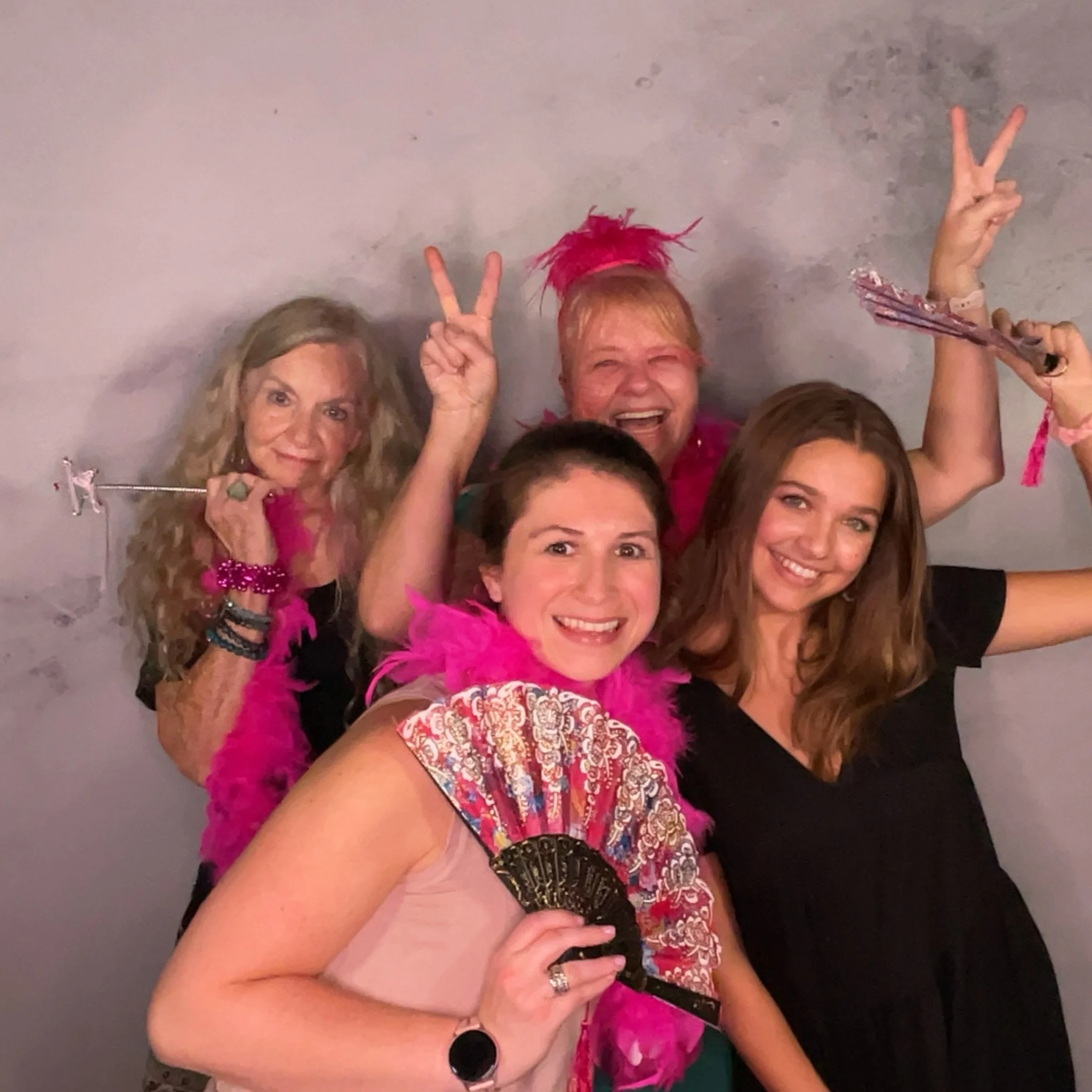 Four women celebrating, smiling, and enjoying themselves, with some holding fans, wearing pink feather boas, and making peace signs.