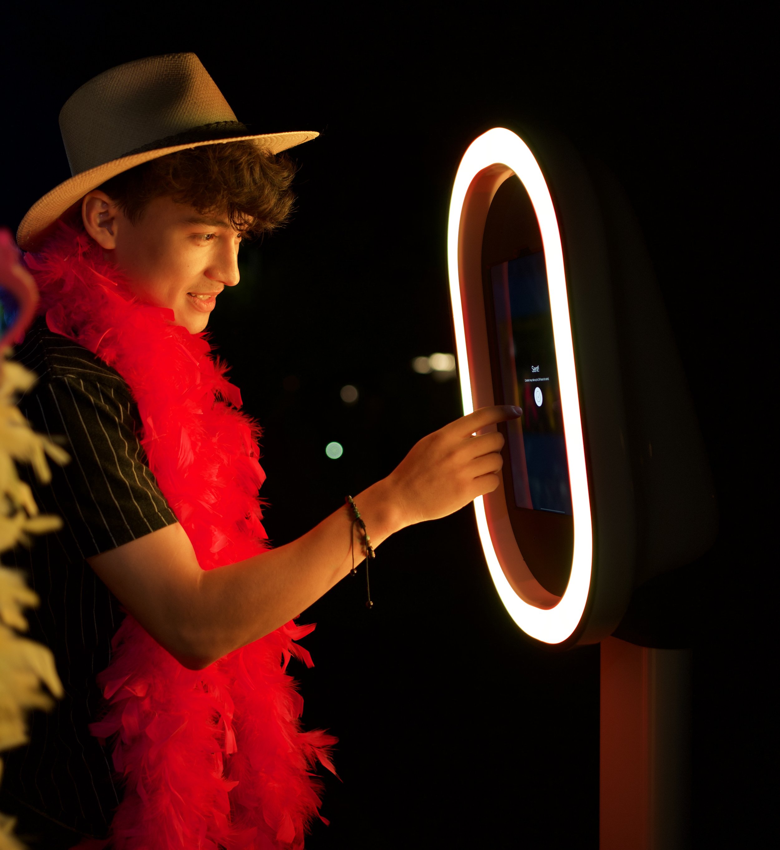 A young man wearing a straw hat, red feather boa, and striped shirt interacts with a futuristic touchscreen kiosk at night, illuminated by a glowing ring around the screen.