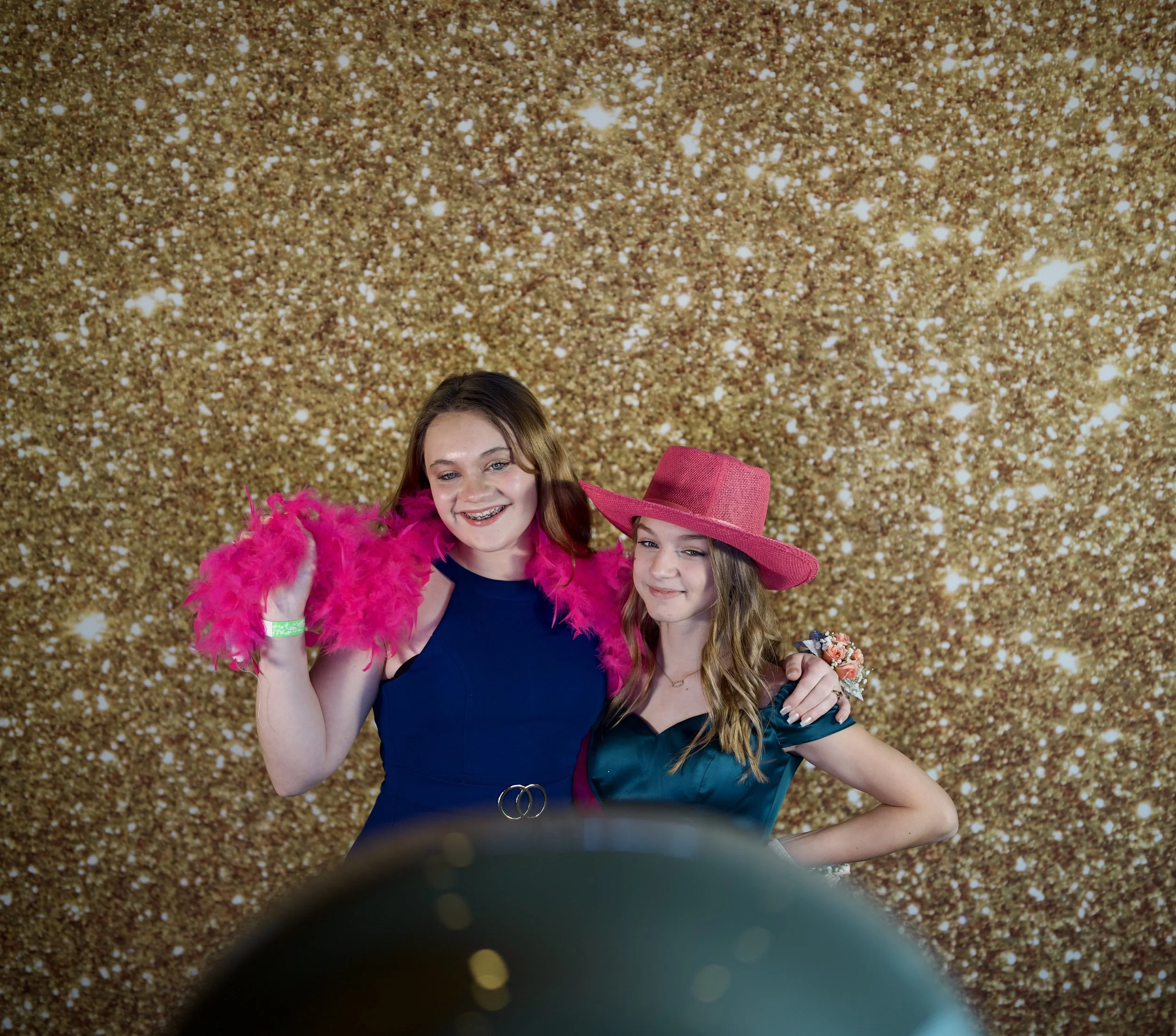 Two young women smiling and posing together in front of a gold glitter backdrop. One wears a pink wide-brimmed hat and a satin dress, the other has long hair and wears a sleeveless navy dress and a bright pink feather boa.