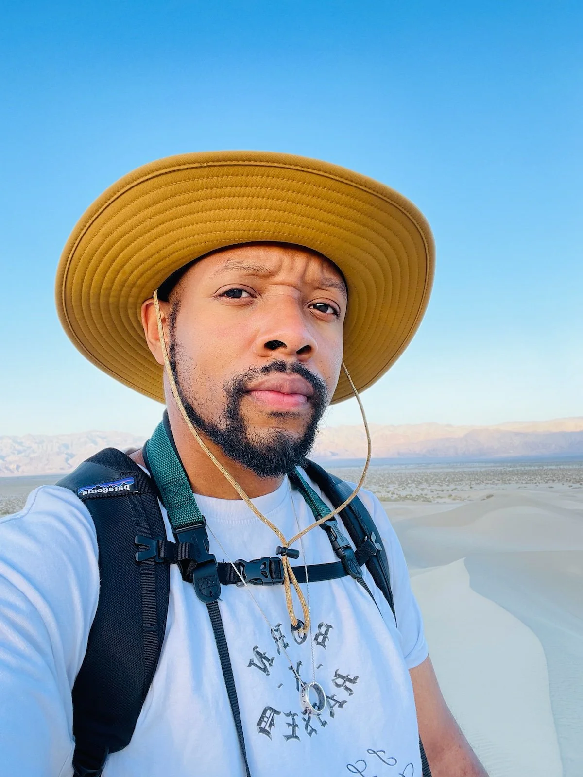 A black man in a tan brimmer hat wearing a silver necklace with a ring attached stands atop beige sand dunes in Death Valley. Mountain ridges far in the background spring to life under the glow of a rising sun.