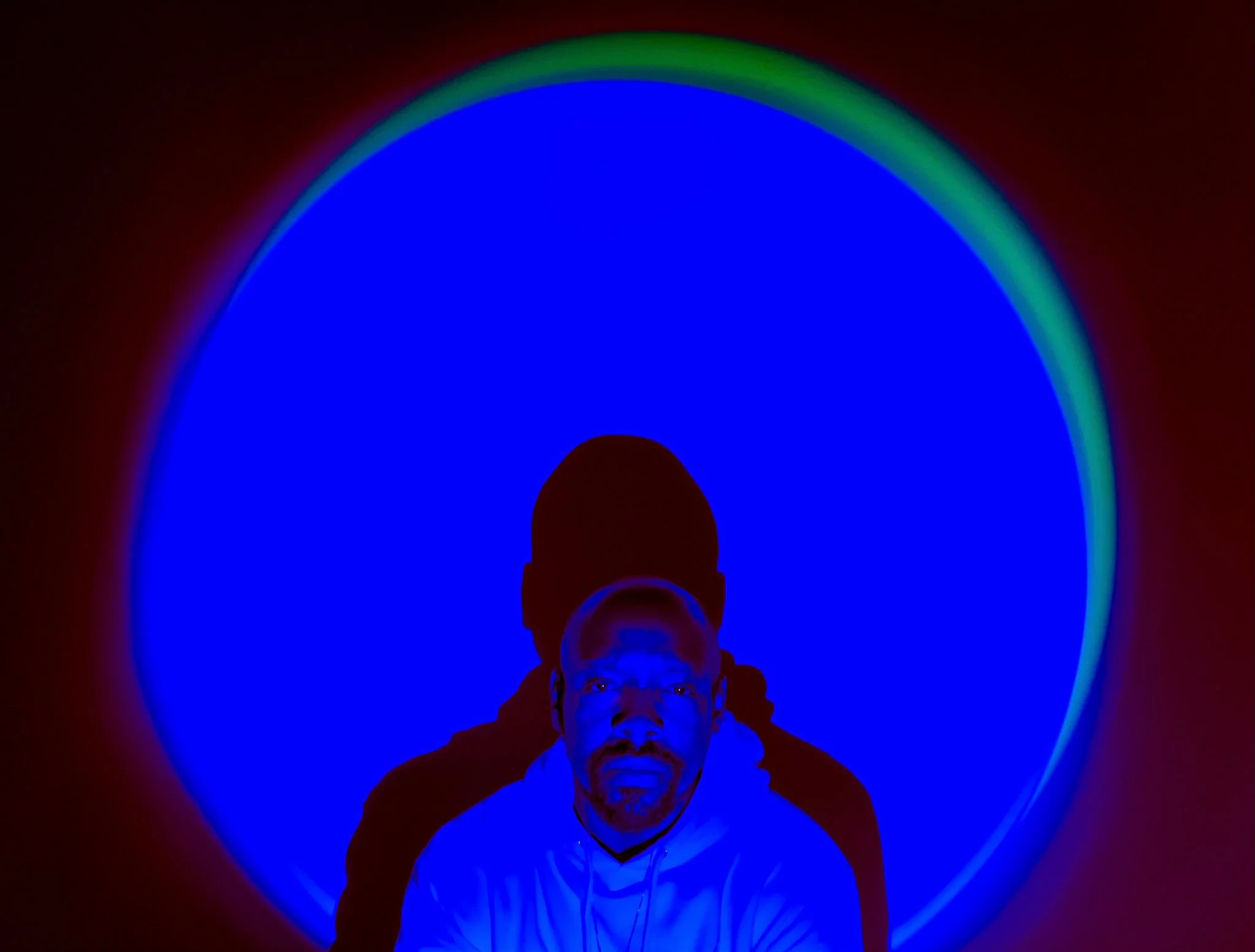 A bald headed black man stares deeply into the lens as neon blue moon of light surrounds his frame.