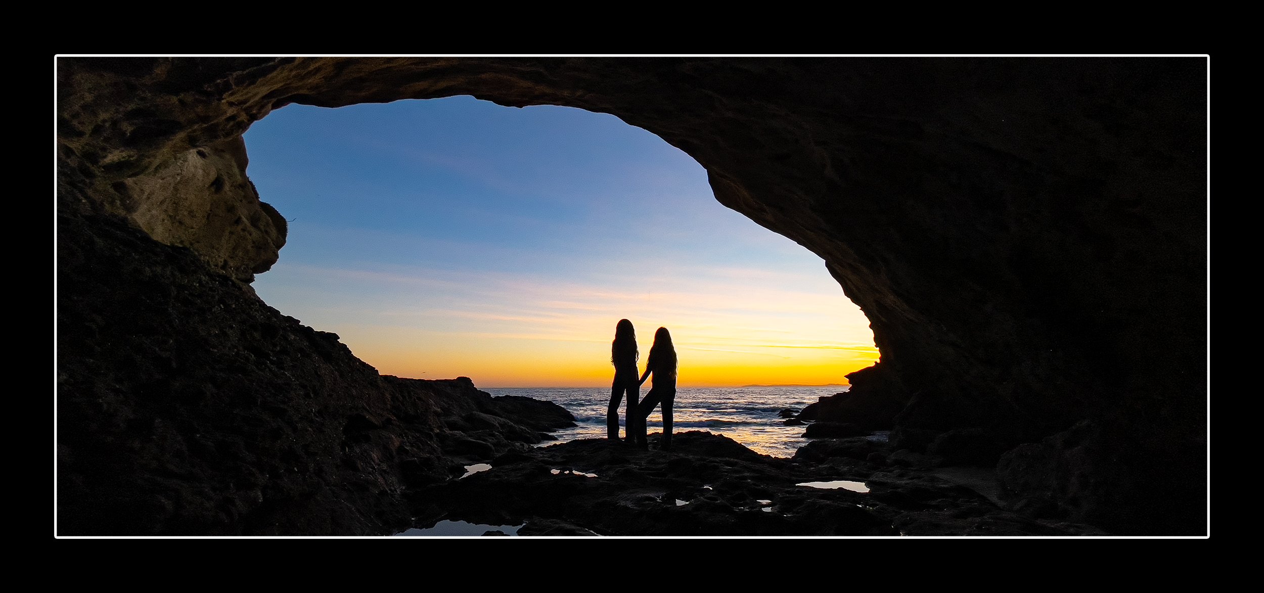 Two girls stand atop a rock bed inside a cave watching the sunset. The orange glow of the sun cuts across the middle of the frame turning the streaking clouds pink as the blue sky fades away.