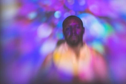 A black man with a thin goatee entrenched in an array of psychedelic colors, vibrant purples, blues, with tiny hits of teal warp around him as he stares deep into the lens.