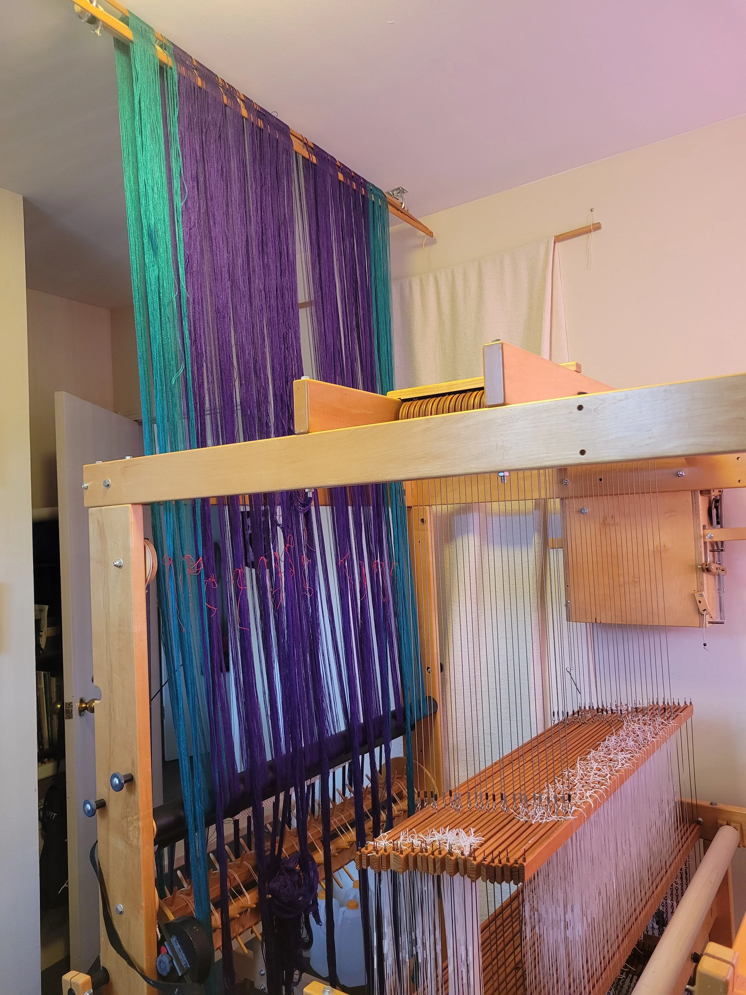 Bouts of warp hung from a "trapeze" to hold them under tension as they get wound into the loom.