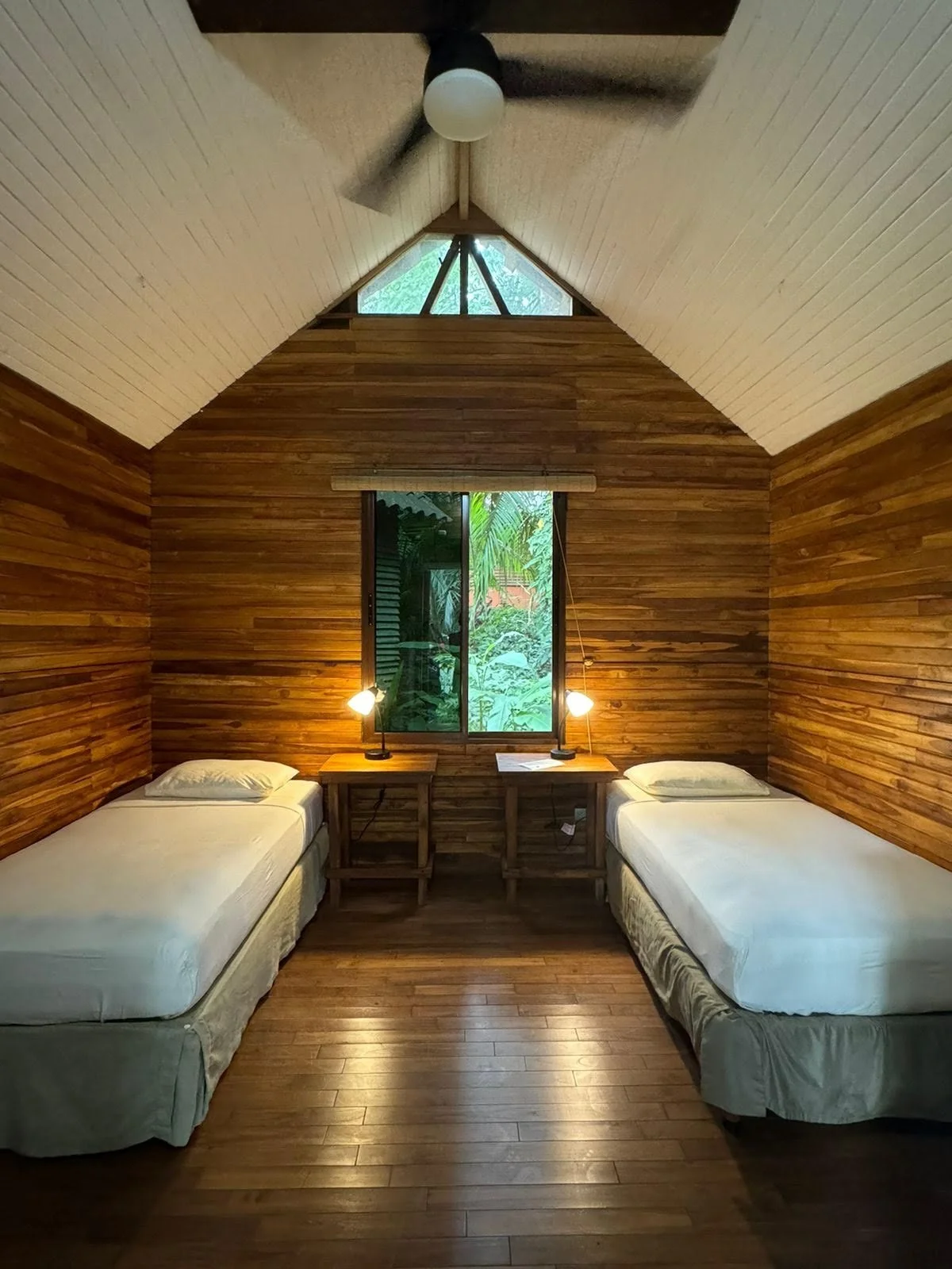 Two small wooden huts surrounded by lush green tropical plants and trees, with stone pathways leading to each hut in a jungle-like setting.
