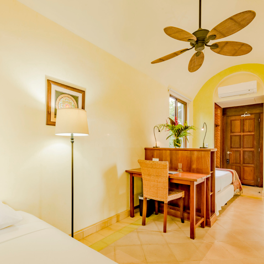 A bedroom with a single bed, wooden headboard, and white bedding, large windows showing trees outside, a ceiling fan, and a small sitting area with a couch and armchair.