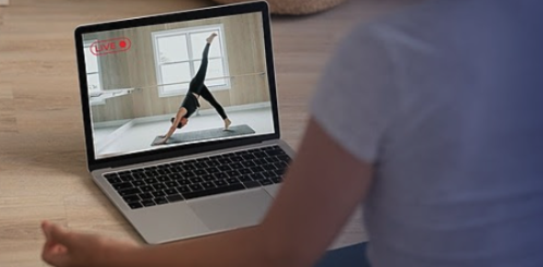 Four women participating in a yoga class in a bright, minimalistic room, practicing downward dog pose with yoga blocks, mats, and water bottles nearby.