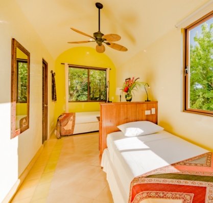 A bedroom with a single bed, wooden headboard, and white bedding, large windows showing trees outside, a ceiling fan, and a small sitting area with a couch and armchair.