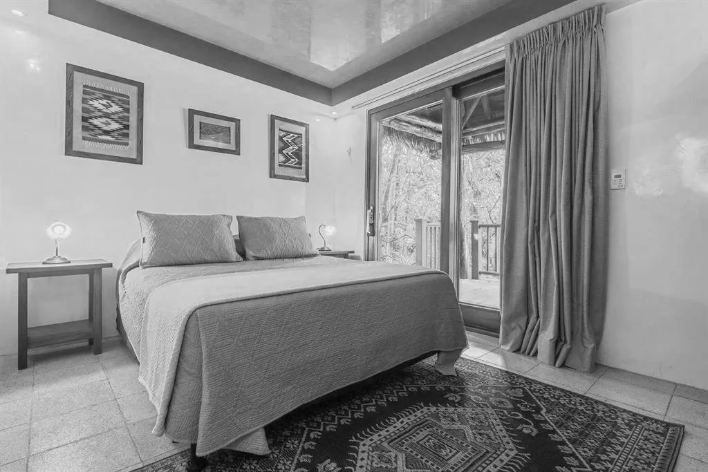 A bedroom with a bed, nightstands with lamps, framed artwork on the wall, sliding glass door with curtains leading to a balcony.