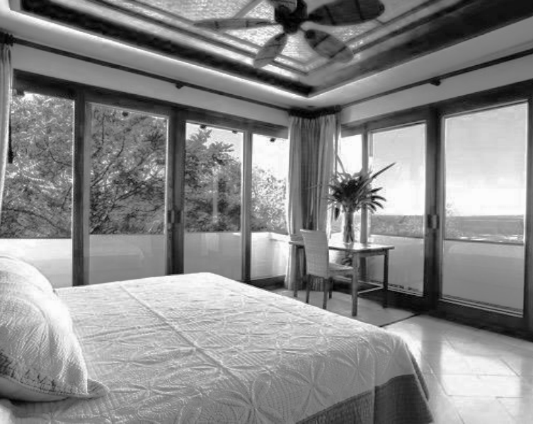 A bedroom with a large bed, glass windows, a small desk with a chair, and a potted plant, overlooking a landscape with trees and open land.
