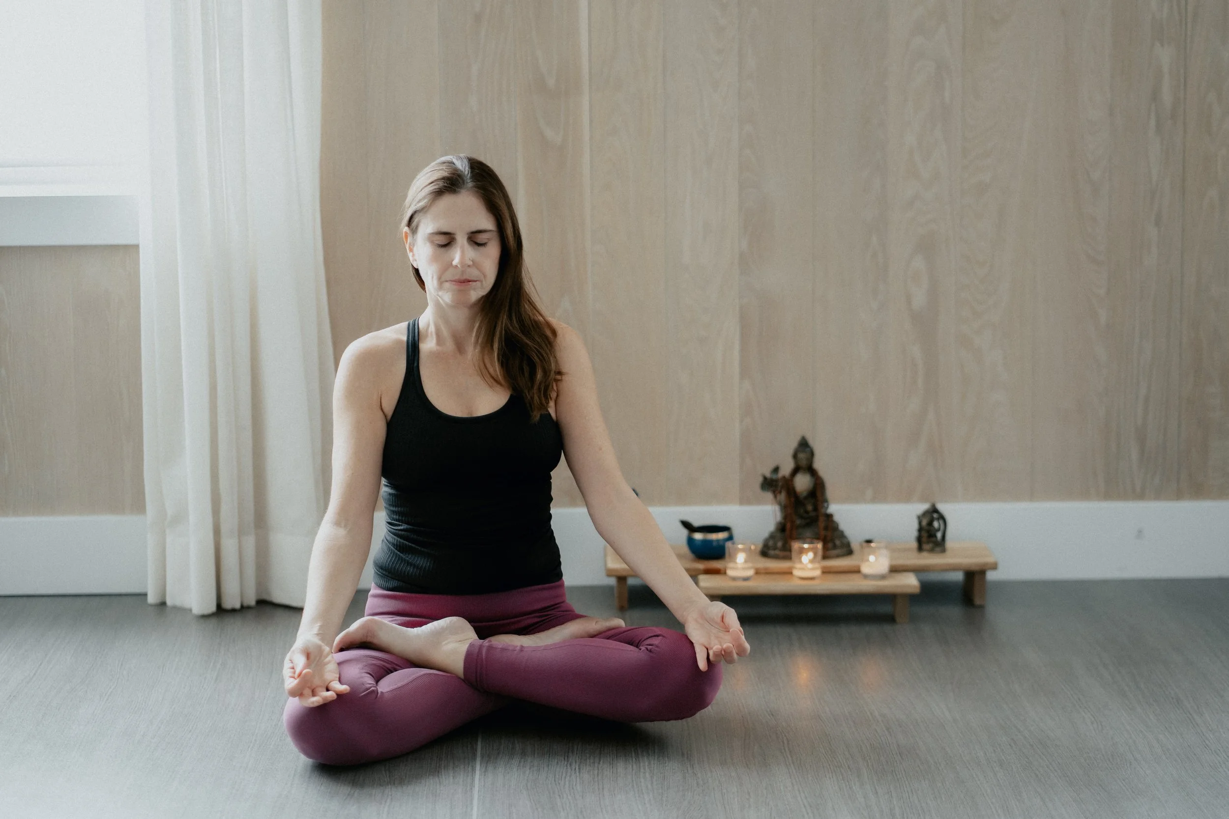 A group of five people participating in a yoga class in a bright room with large windows and white curtains, sitting on yoga mats with arms extended outward, led by an instructor sitting cross-legged at the front.