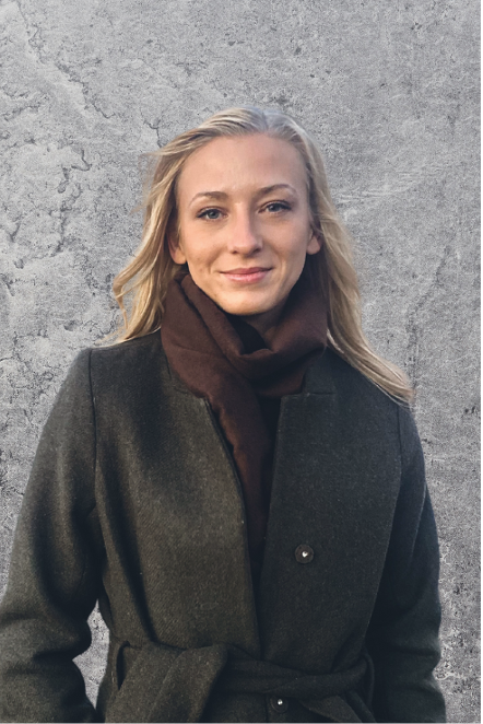Bild på Dominika utomhus, som i en mörk kappa blickar in i kameran med en grå vägg i bakgrunden