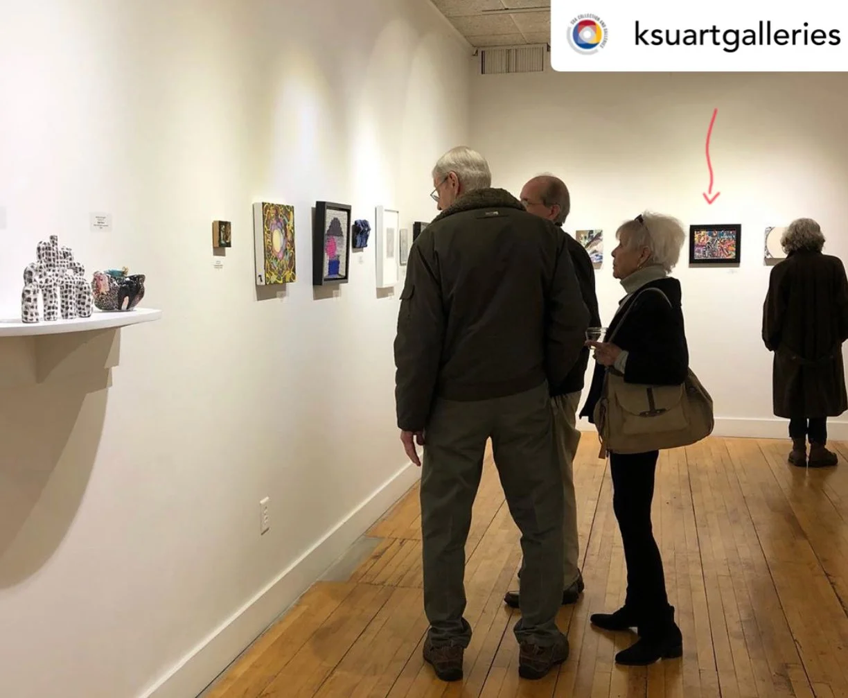 People viewing artwork in an art gallery with framed pieces on the wall and pottery on a shelf to the left.
