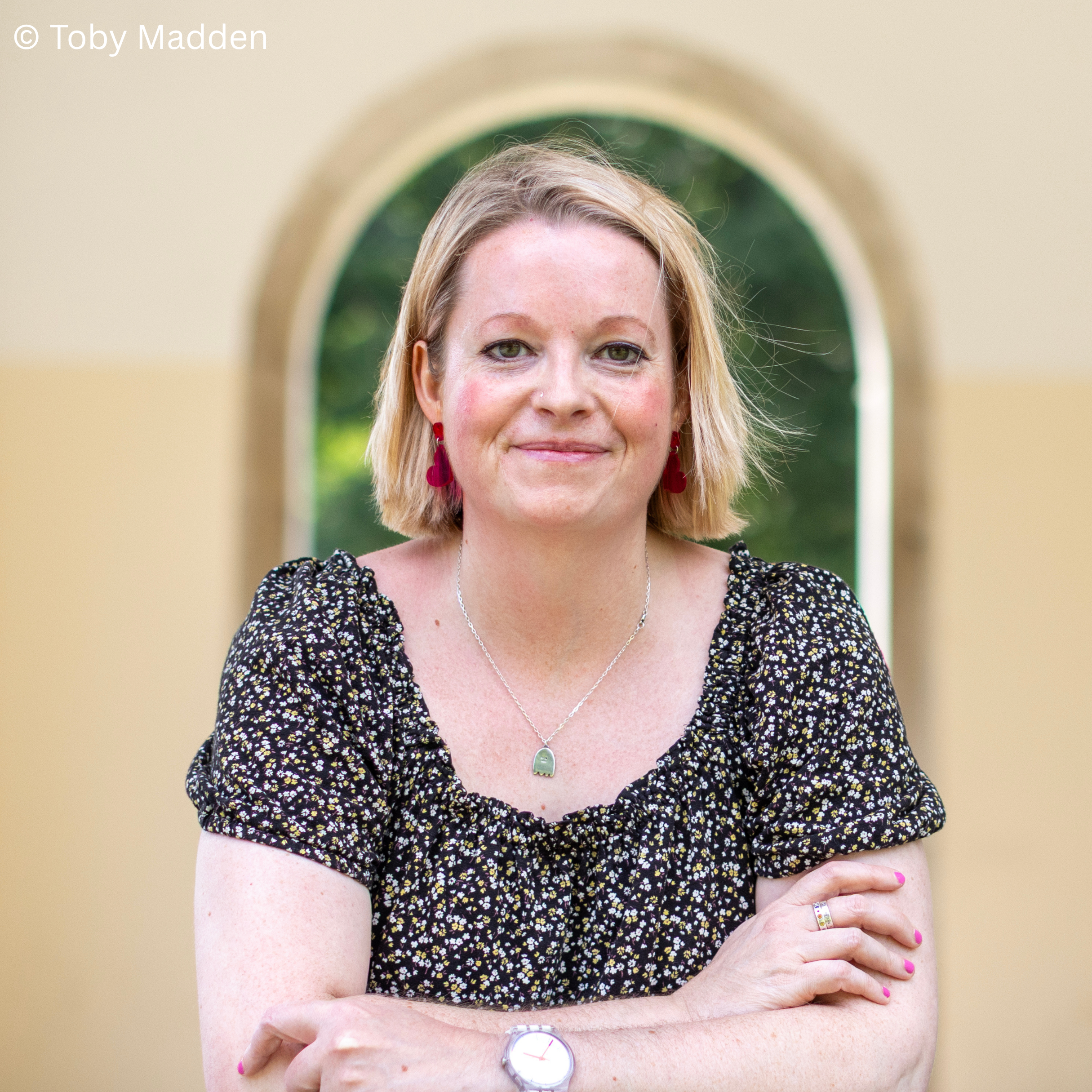 Cressida McLaughlin with blonde hair and green eyes, smiling softly, wearing a black floral top and a ghost pendant, a white wrist watch, standing in the sunshine, crossing her arms in front of a yellow wall with an arched mirror reflecting greenery.