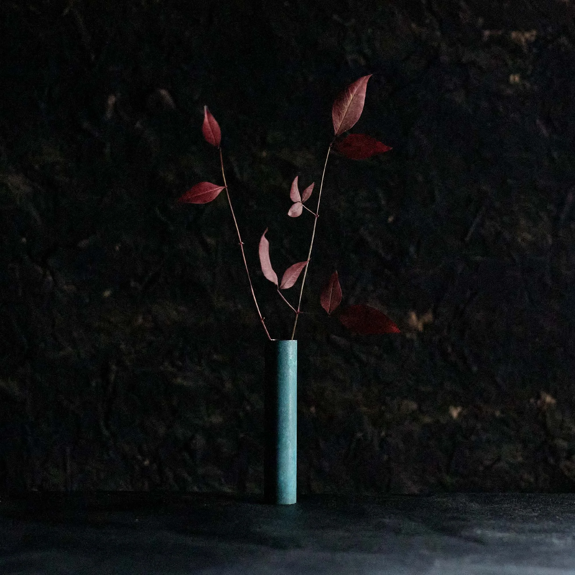 葉のついた細い枝を入れた青色の長い筒が置かれている写真。背景は暗く見える。
