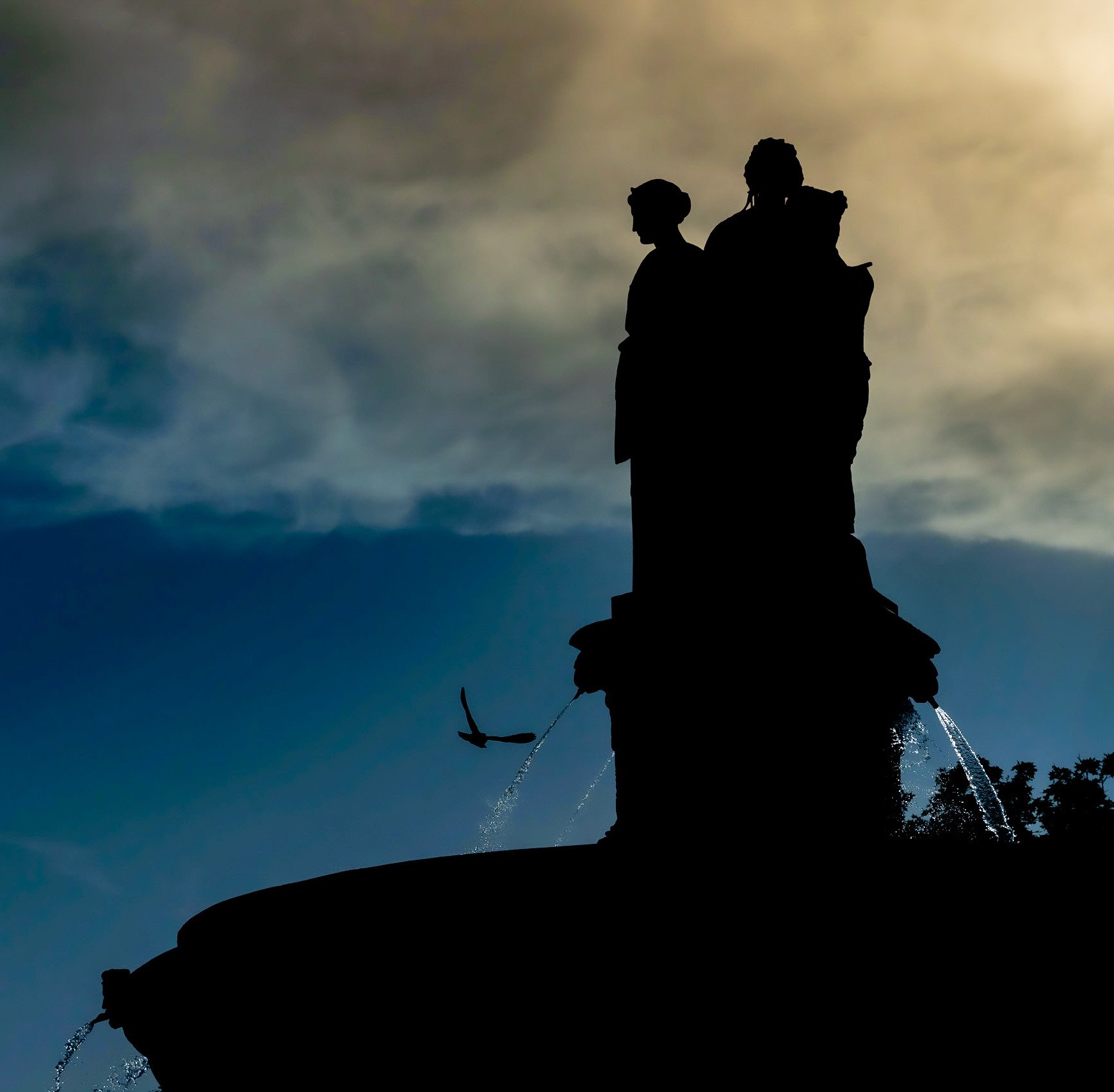 Silhouette de statues sur une fontaine avec un ciel nuageux en arrière-plan, et un oiseau en vol.