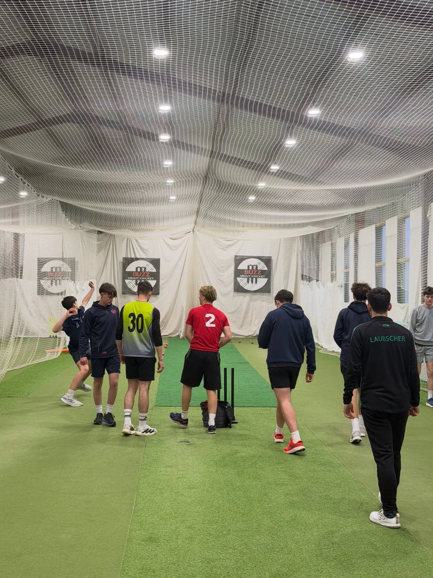 Group of young soccer players and coach inside an indoor training facility with green artificial turf, white netting, and banners on the walls.