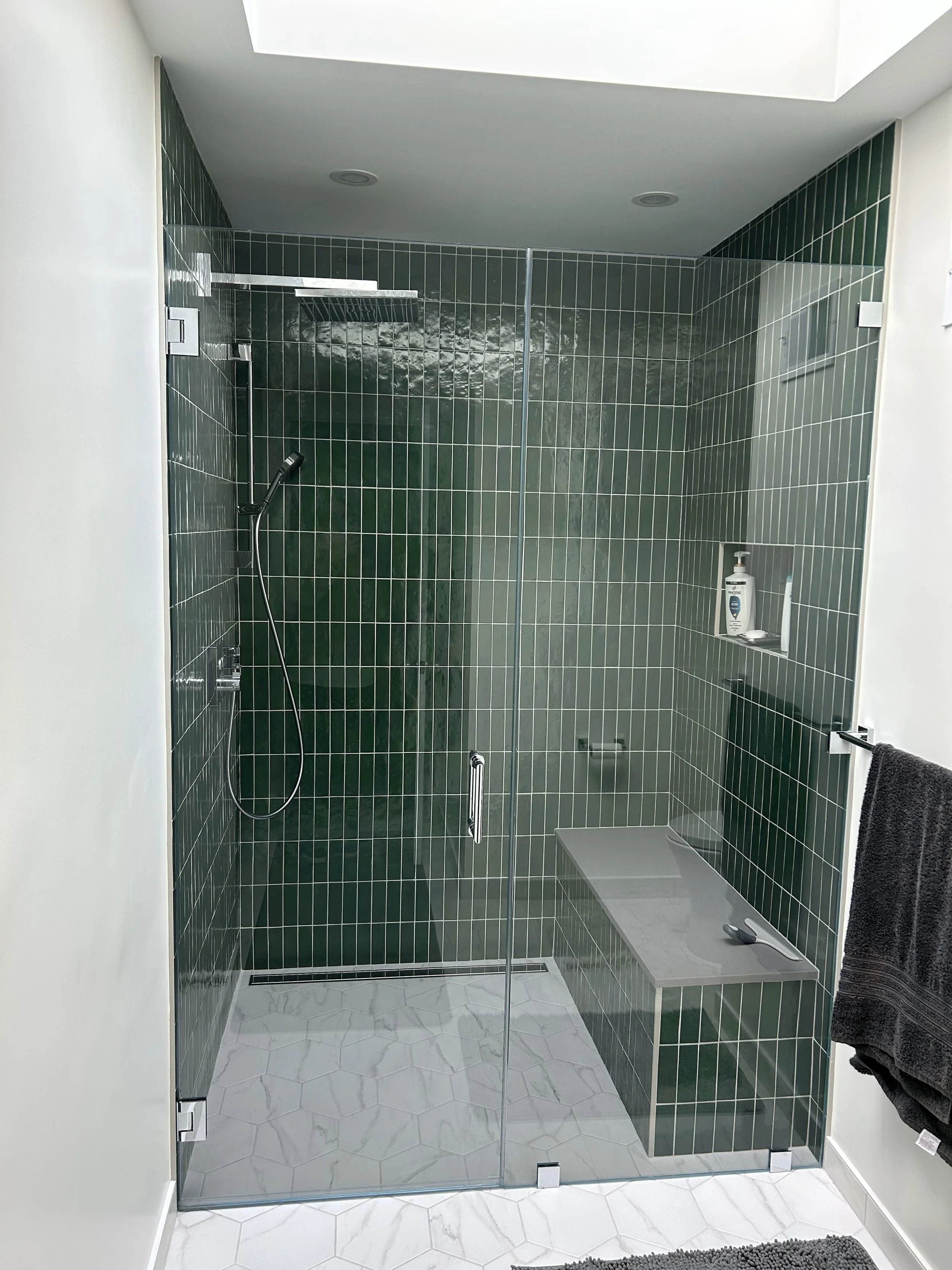A modern walk-in shower with a glass door, green tiled walls, a wall-mounted showerhead, and a built-in bench with a spoon and a towel hanging on a nearby towel bar.