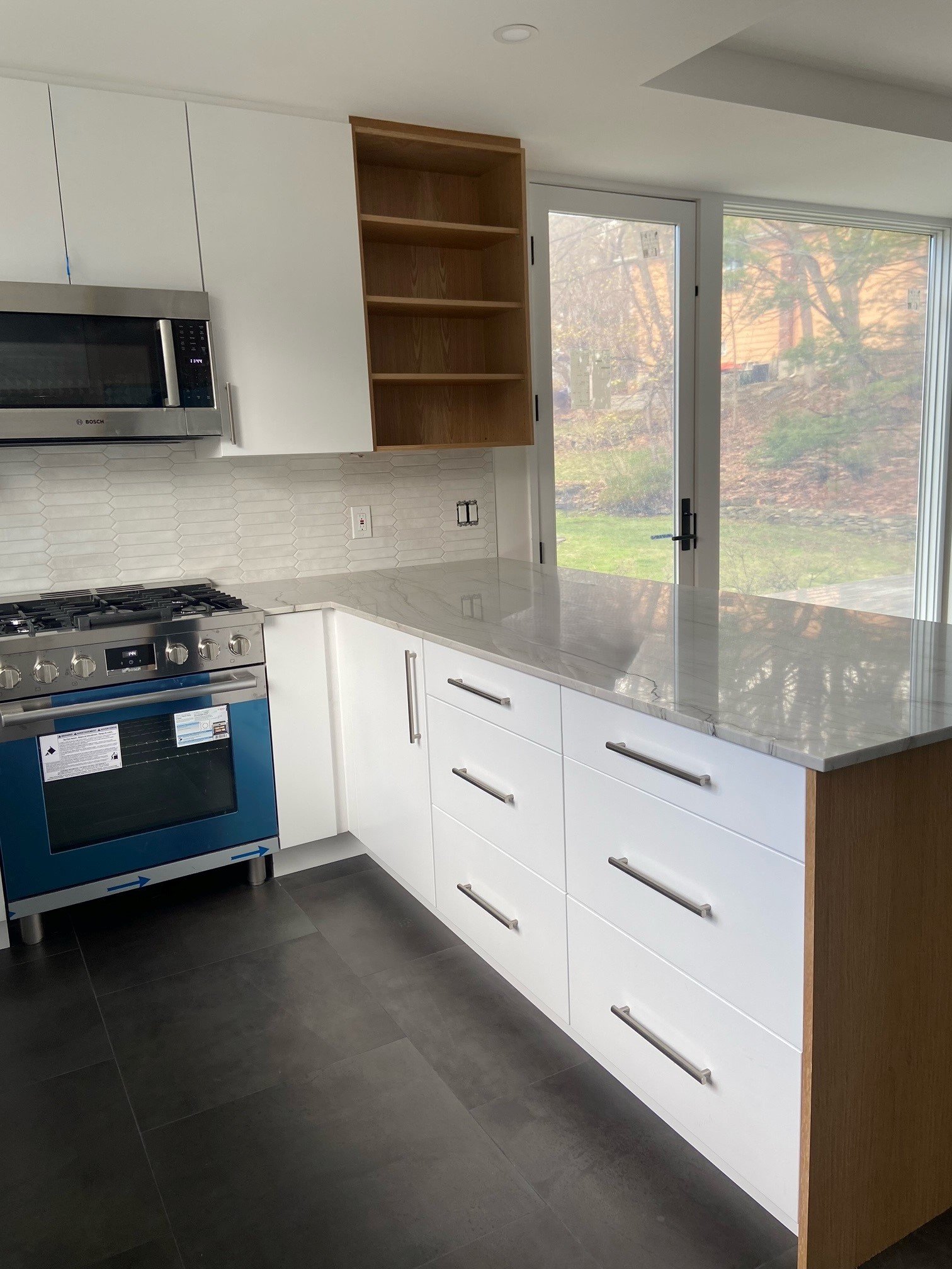 Modern kitchen with white cabinets, a marble countertop, built-in oven, microwave, open wooden shelves, and large windows showing an outdoor view.