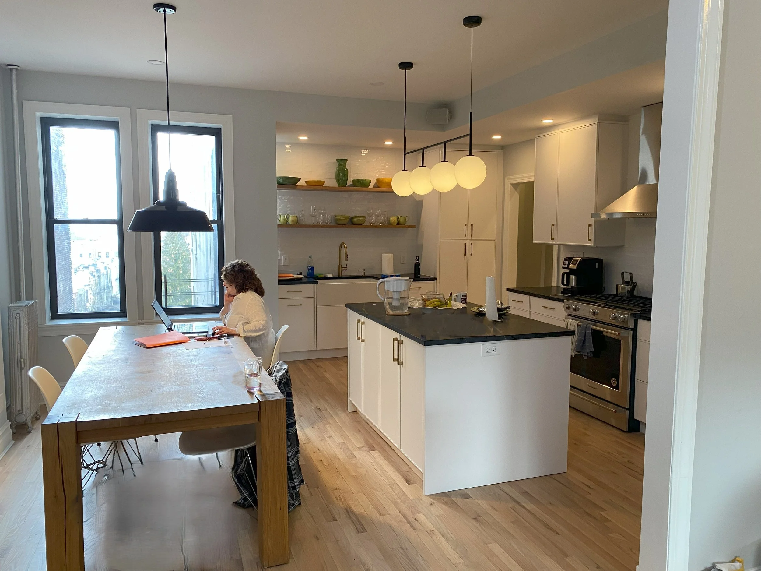 A woman sitting at a dining table working on a laptop in a modern kitchen with white cabinets, black countertops, and three windows, with sunlight coming through.