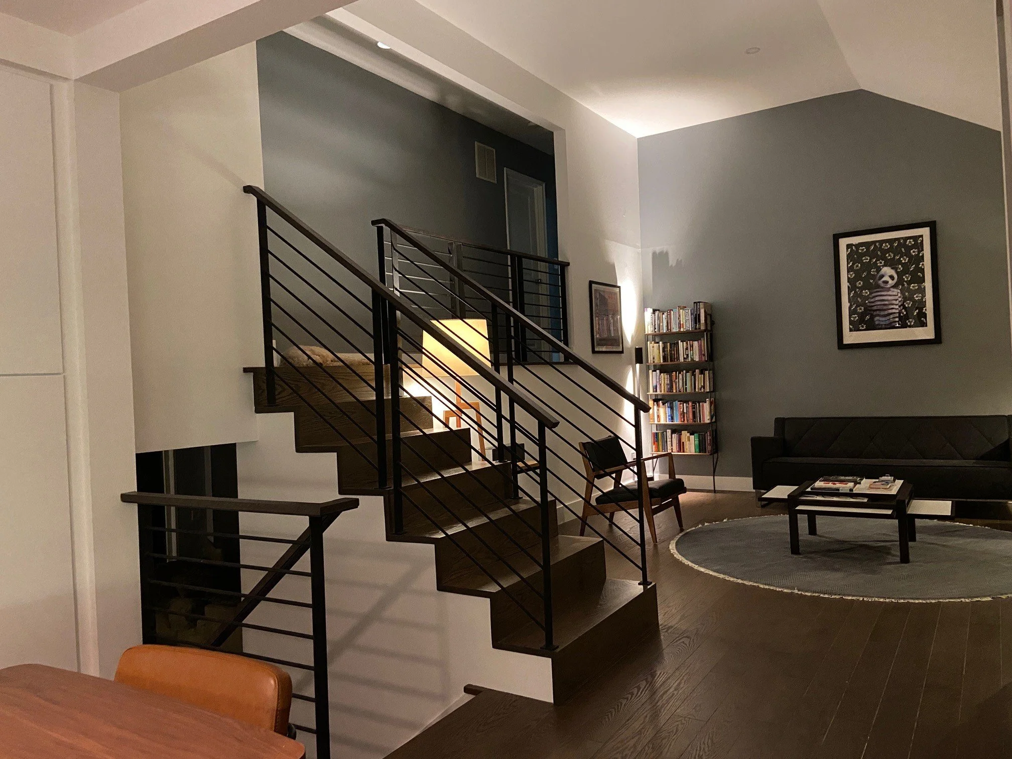 A living room with a staircase, a bookshelf, a black sofa, a round rug, a coffee table with magazines, a chair, and framed artwork on the wall.