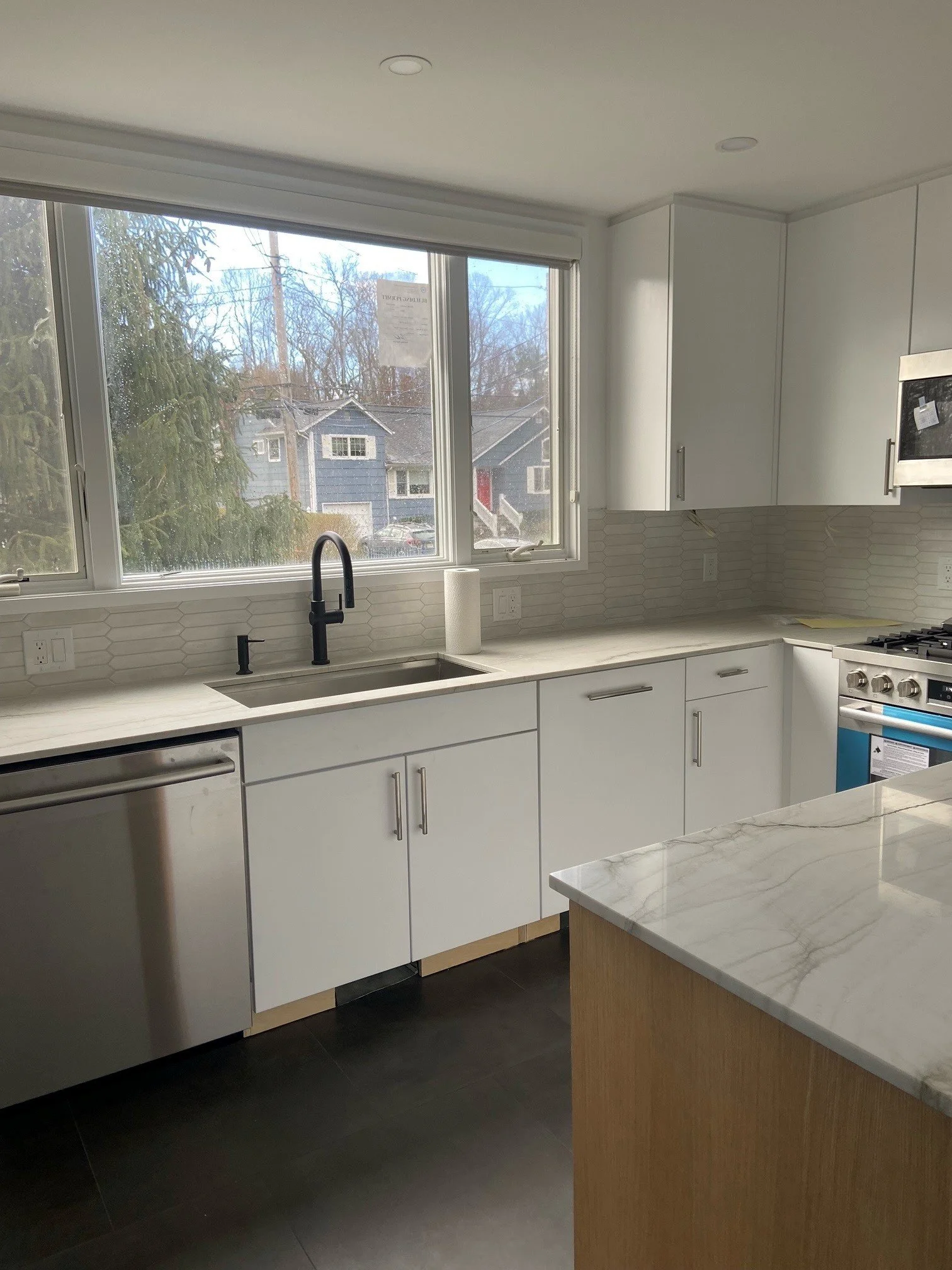 Modern kitchen with white cabinets, marble countertop island, black faucet, and large window overlooking suburban neighborhood.