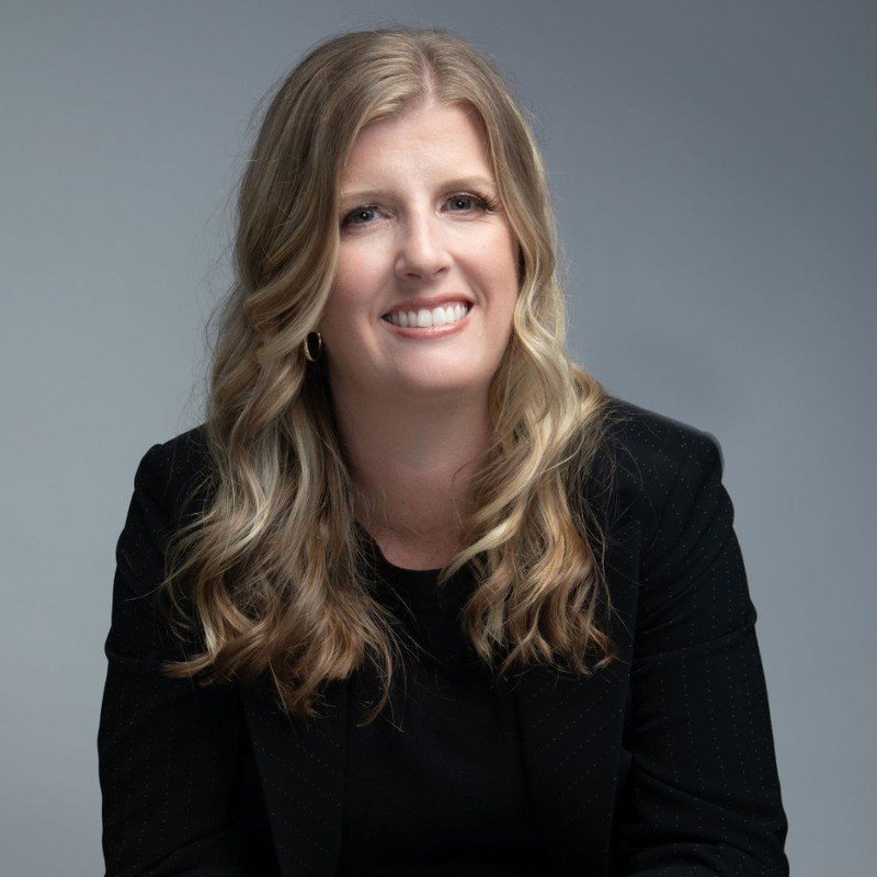 A woman with long, wavy blonde hair and light skin, smiling and wearing a black blazer, sitting against a gray background.