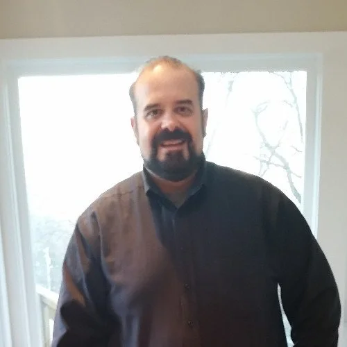 A man with a beard and mustache wearing a dark button-up shirt standing indoors near a window with trees outside.