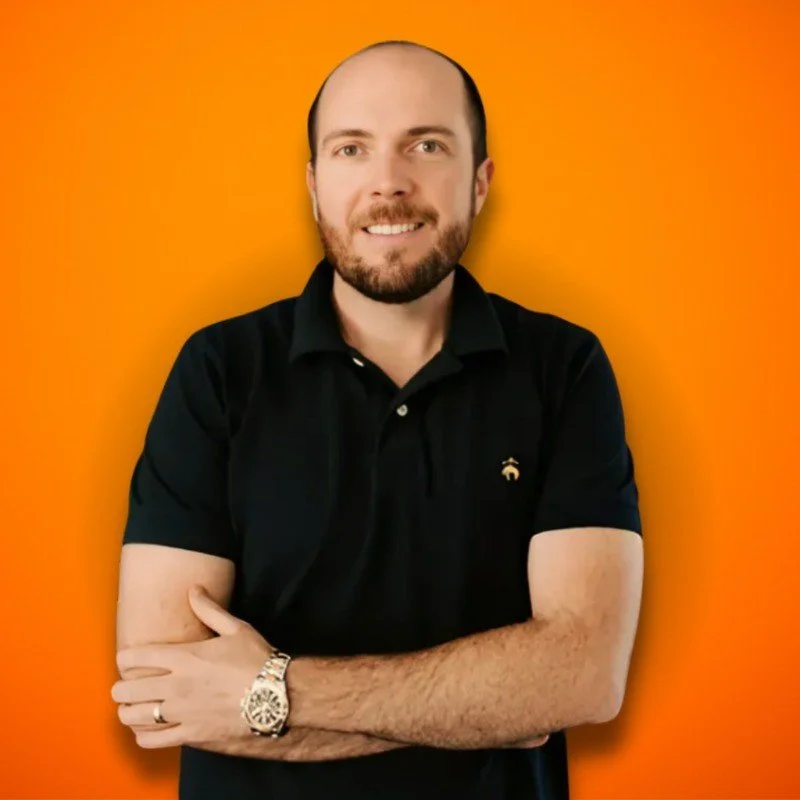 Photo of a smiling man with a beard and bald head, wearing a black polo shirt, standing against an orange background.