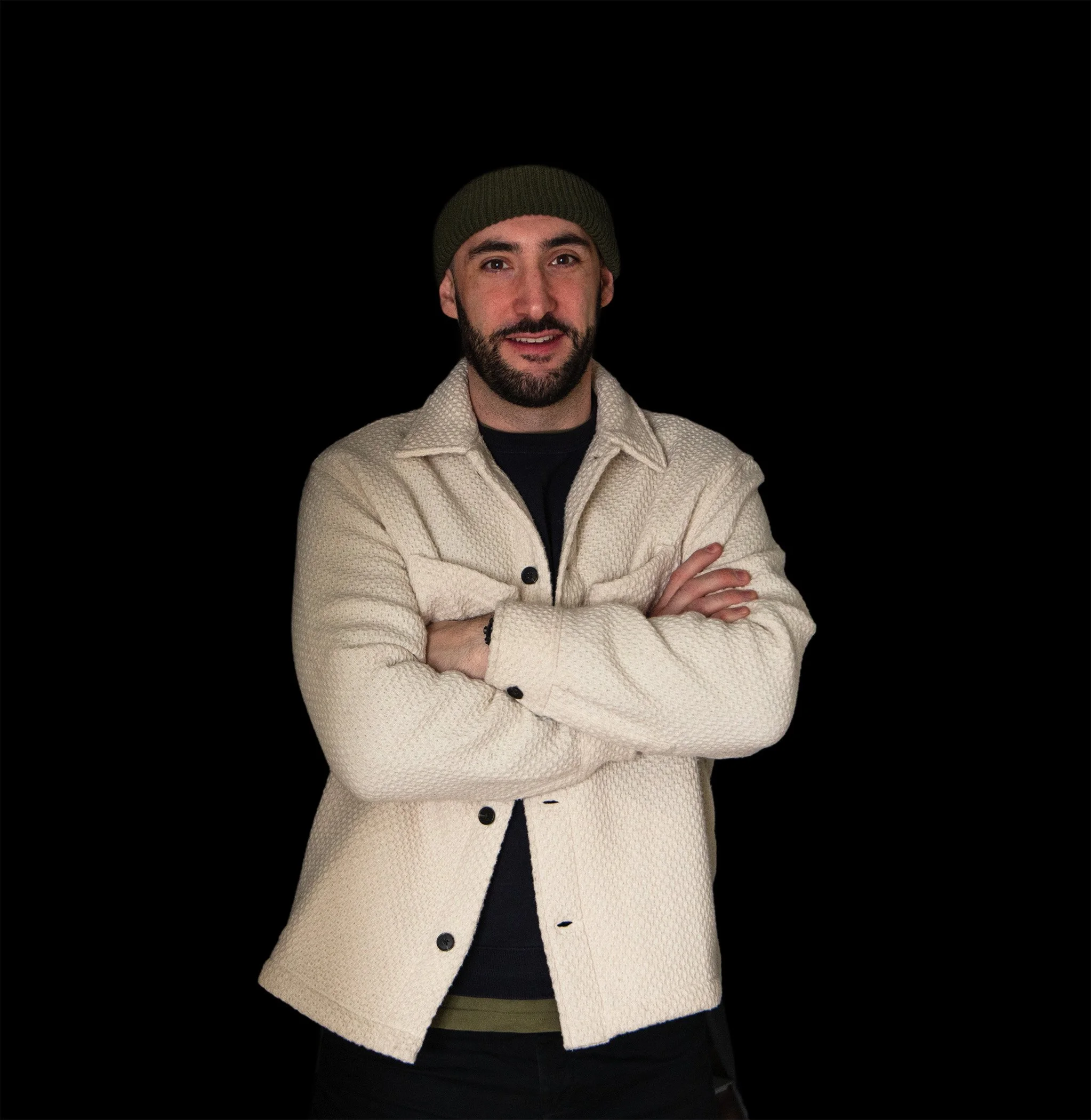 Un homme avec une barbe porte un bonnet vert et un surchemise blanche, croisant les bras, sur un fond noir.