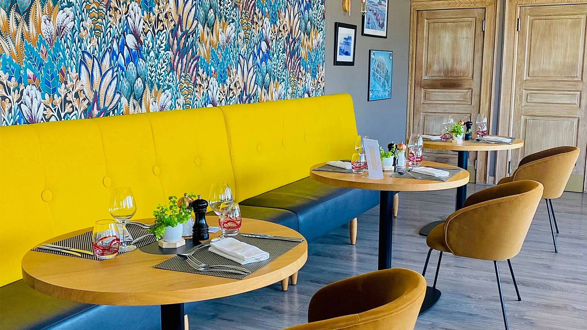 Salle à manger dans un restaurant avec banquette jaune, chaises beige, tables en bois, décoration florale, verre et set de table, murs en bois et mur décoré de papier peint coloré à motifs végétaux.