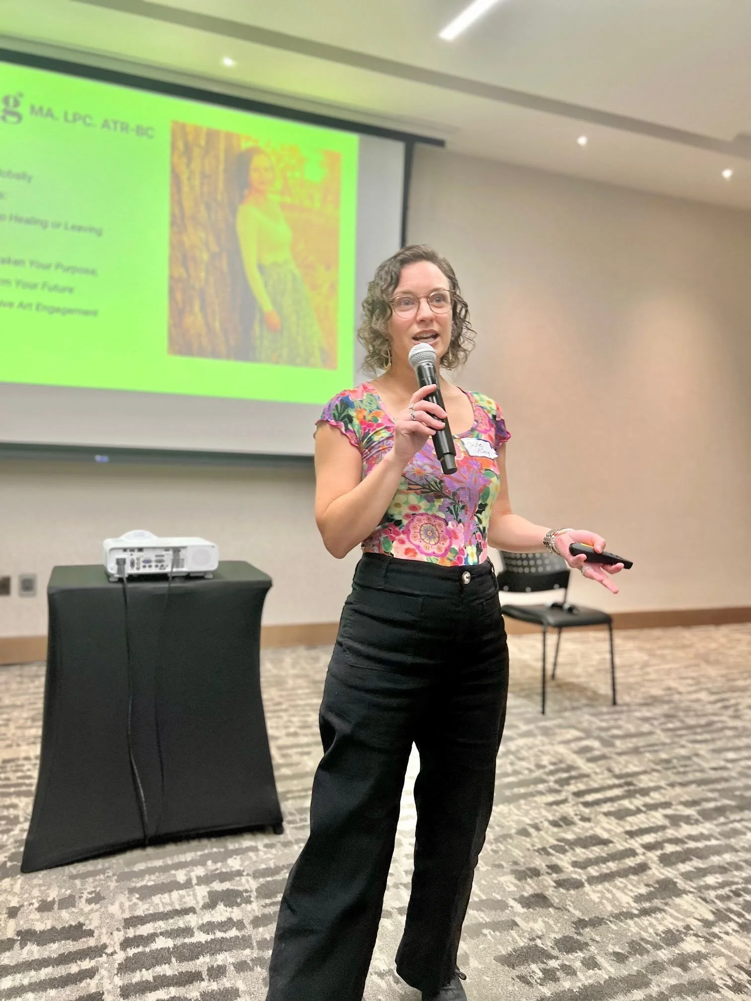 A woman with curly hair and glasses speaking into a microphone during a presentation in a conference room. She is wearing a colorful floral top and black pants. A presentation slide with a picture of her and some text is projected on the wall behind her.