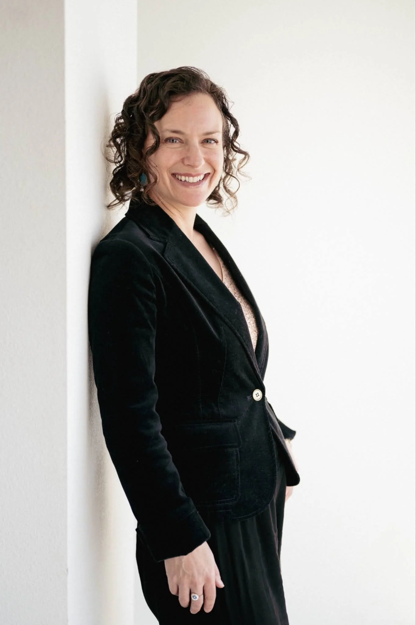 A woman with curly brown hair smiling and leaning against a white wall. She is wearing a black blazer and a ring with a large stone.