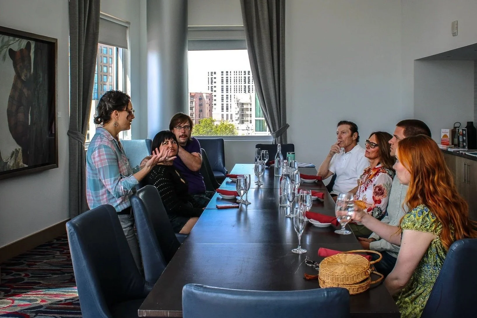 People gathered around a long dinner table in a high-rise apartment, with a woman standing and speaking to the group, while others listen attentively. The table is set with glasses, red napkins, and some personal items.