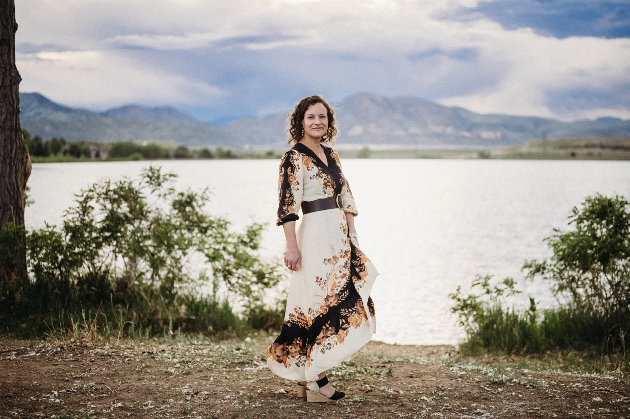Woman standing near a lake with mountains in the background, trees, and cloudy sky.
