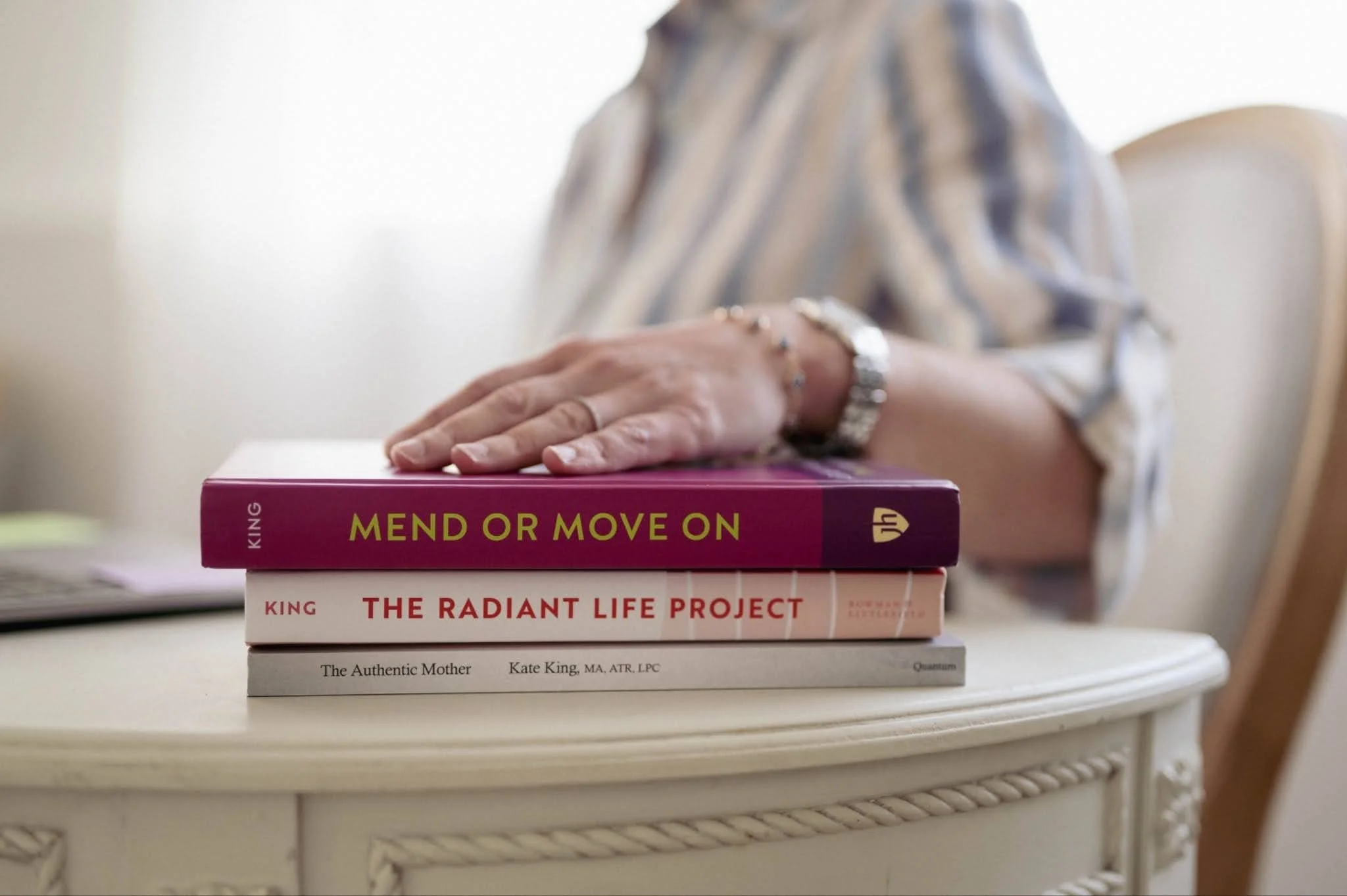 A woman wearing a striped shirt leaning on a stack of three books on a white table. The books are titled 'Mend or Move On', 'The Radiant Life Project', and 'The Authentic Mother'. The woman's face is not visible.