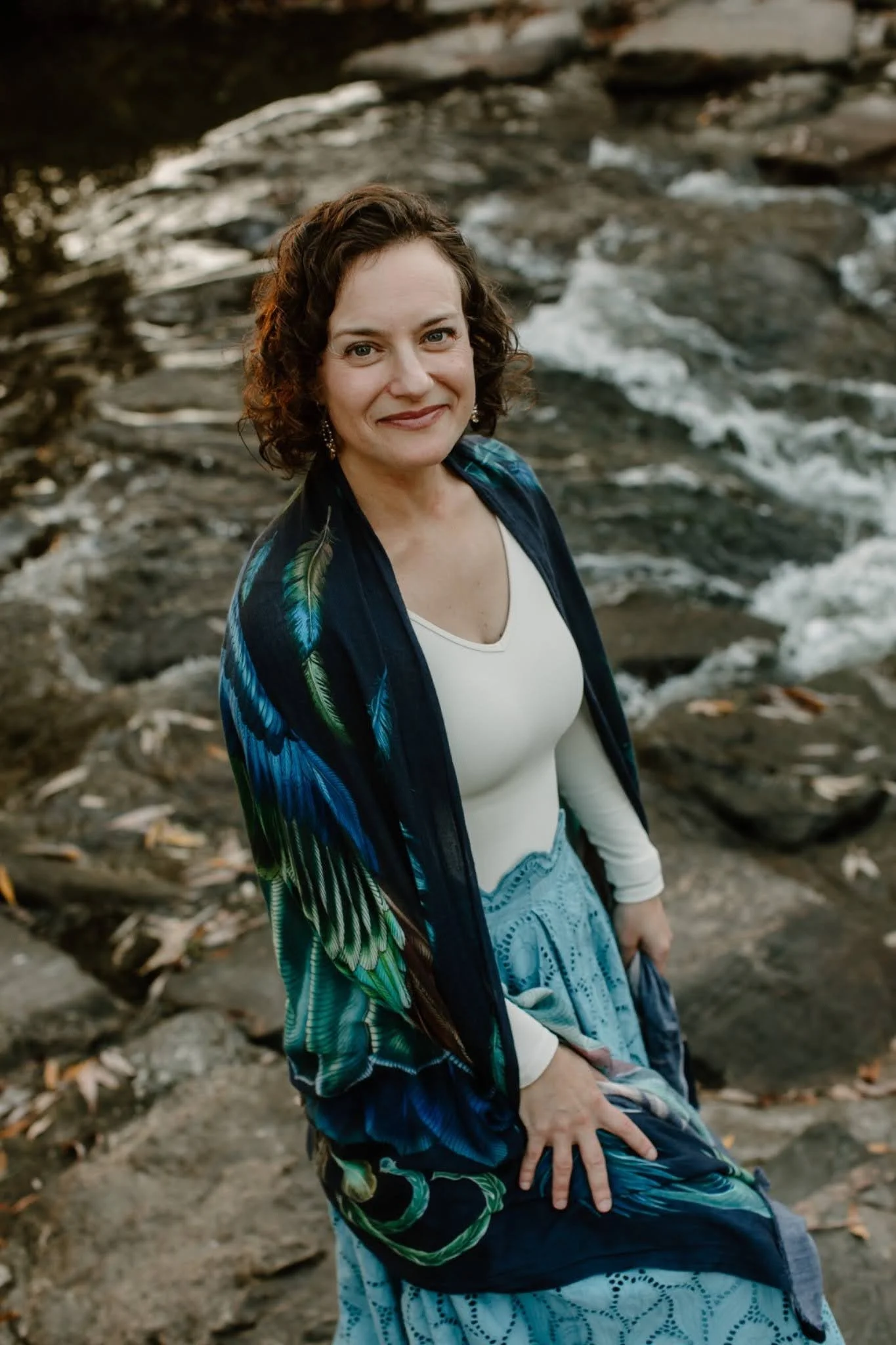 A woman with curly brown hair and earrings standing near a rocky, flowing river or creek, smiling at the camera.