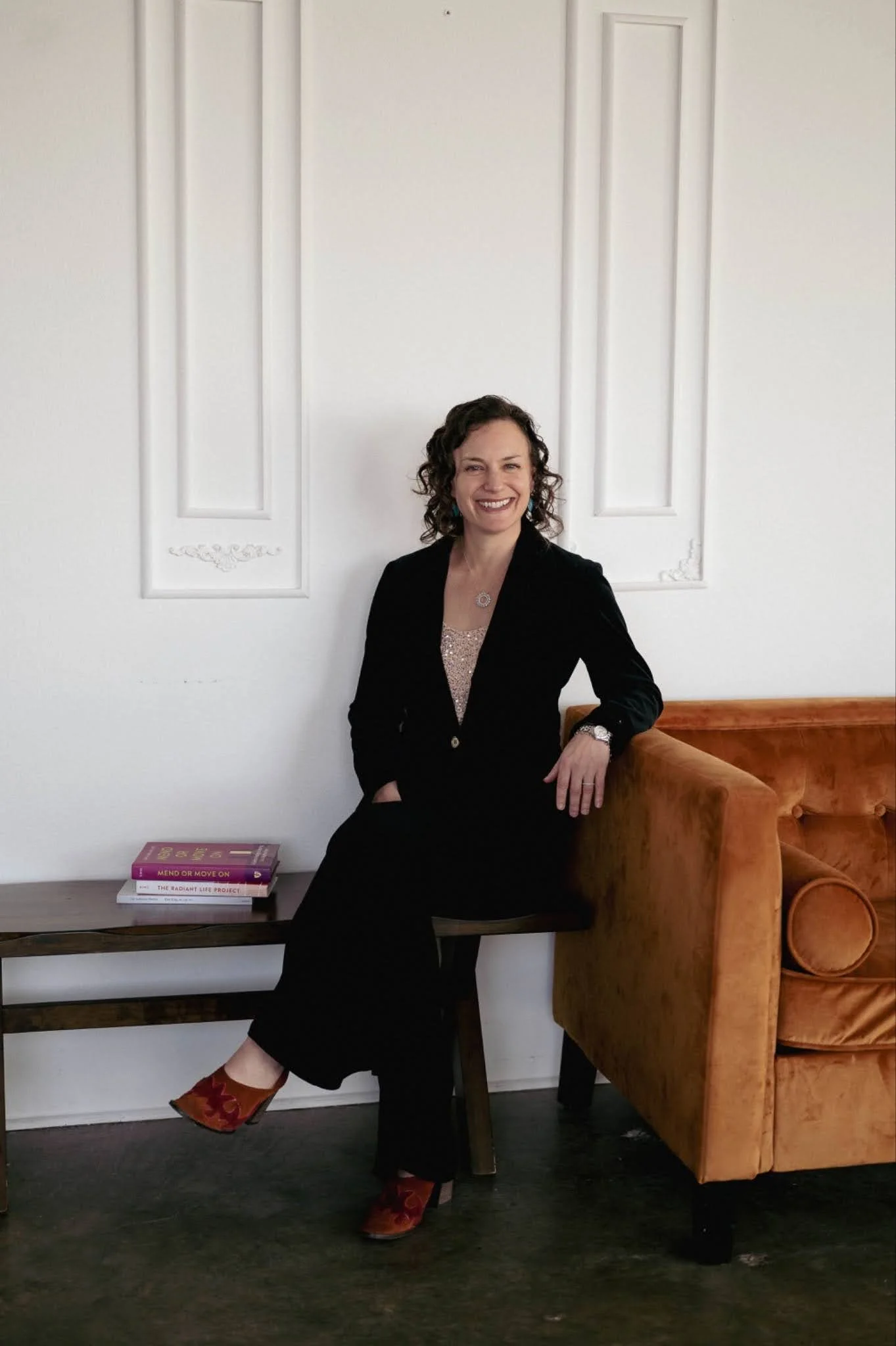 A woman with curly dark hair and a bright smile, wearing a black blazer and pants, seated on a wooden bench next to a brown velvet armchair. She is wearing orange and red patterned shoes and turquoise earrings. Behind her is a white wall with decorative molding. On a small table beside her, there are three books stacked, titled "Mend or Move On," "The Radiant Life Project," and "The Future of Work."