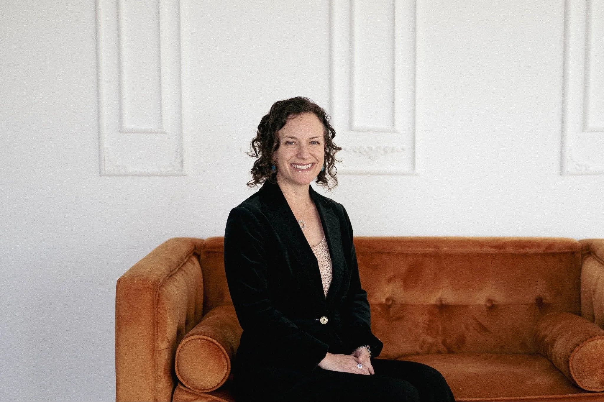 A woman with curly dark hair, wearing a black blazer, sitting on an orange velvet couch in a white room with ornate wall decor, smiling at the camera.